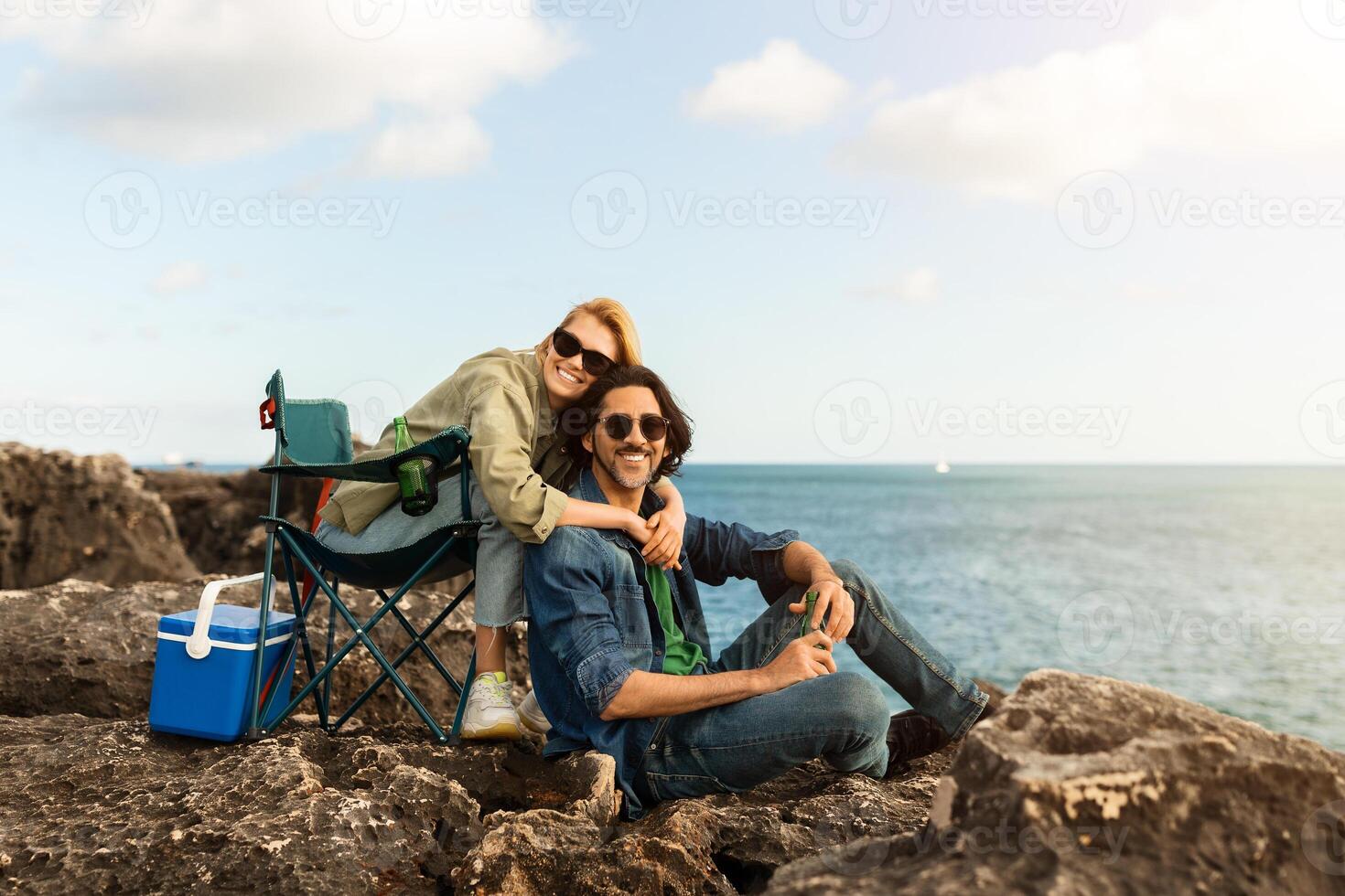A couple relaxes on the rocky beach, with the woman playfully embracing the man. They enjoy their picnic, surrounded by water and a clear blue sky on a bright day. photo