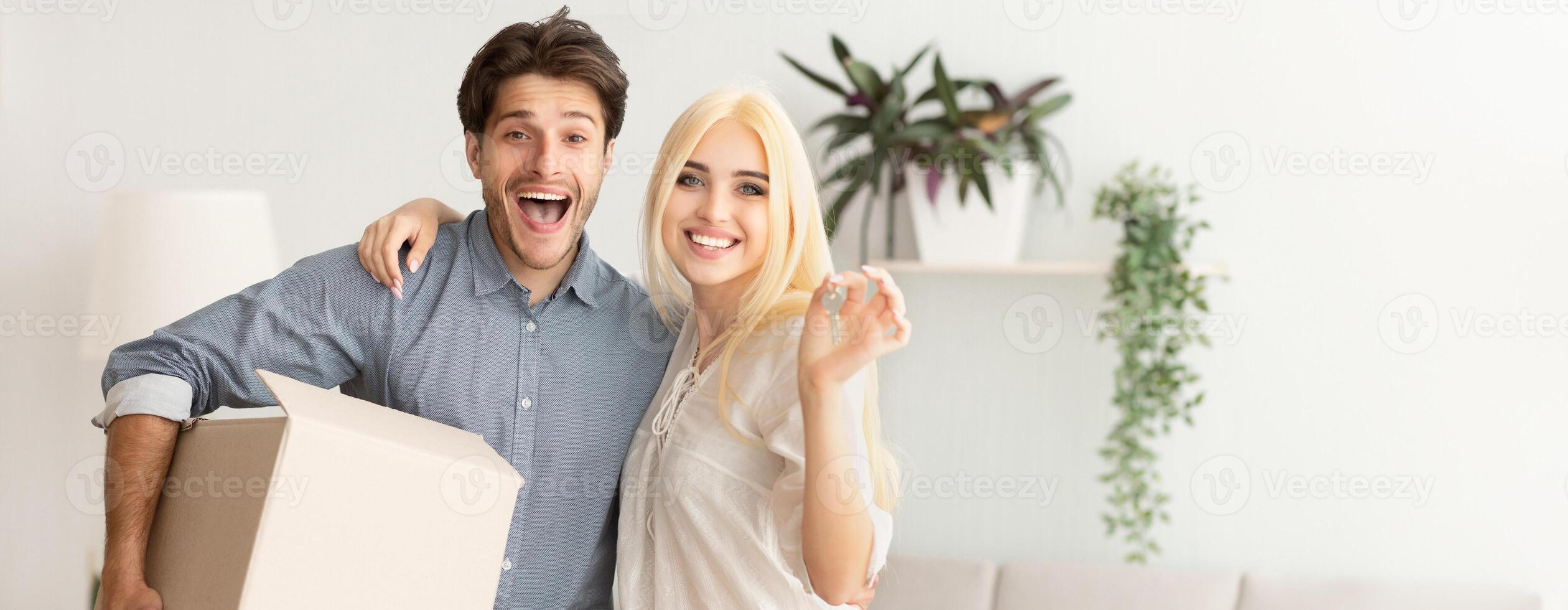 A joyful couple poses in their new living room, holding a cardboard box and smiling broadly. They are surrounded by plants and a bright, welcoming atmosphere, indicating a fresh start, copy space photo
