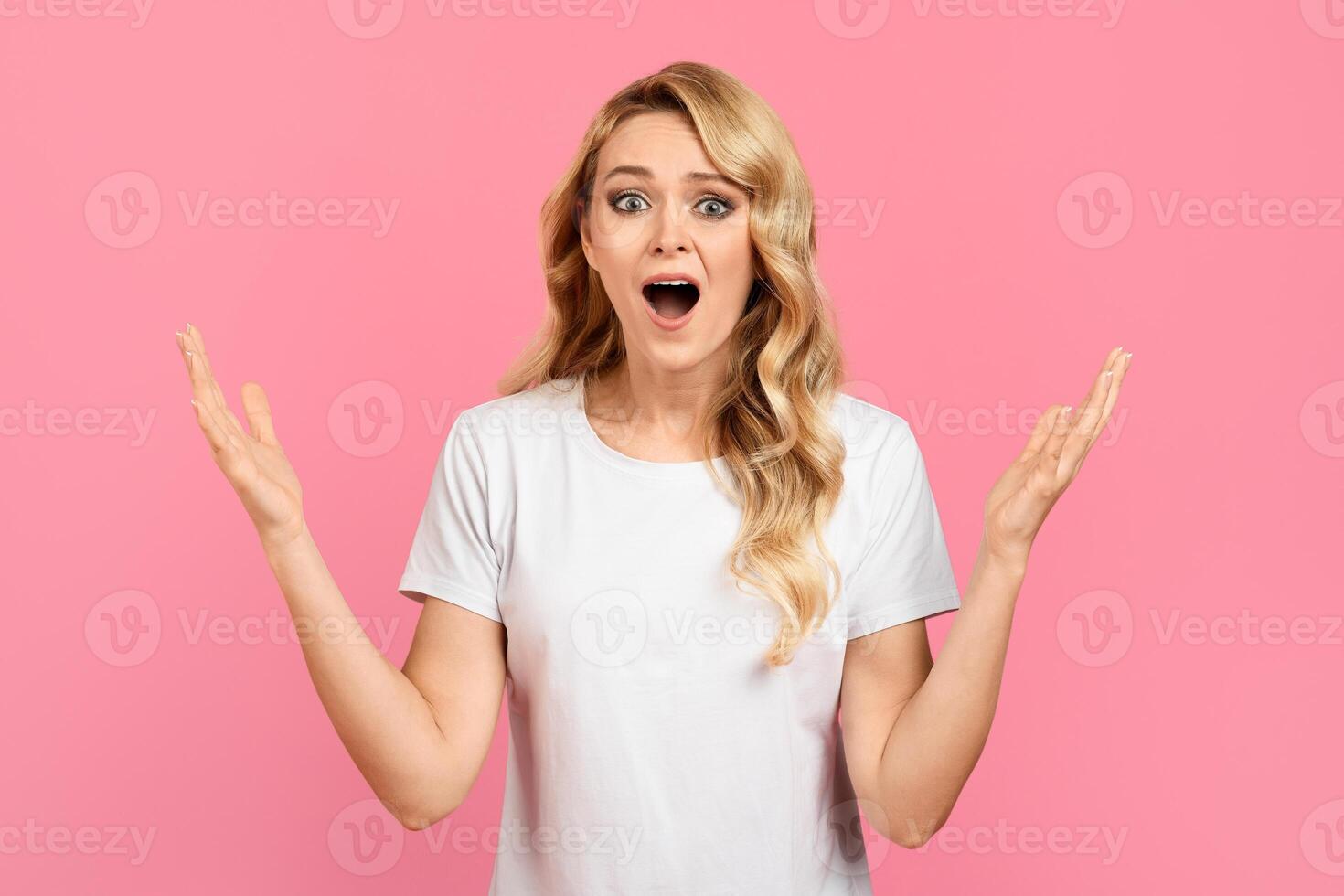 A woman with long blonde hair is standing against a pink background. She has an astonished expression, mouth open, and arms raised in a gesture of surprise and excitement. photo