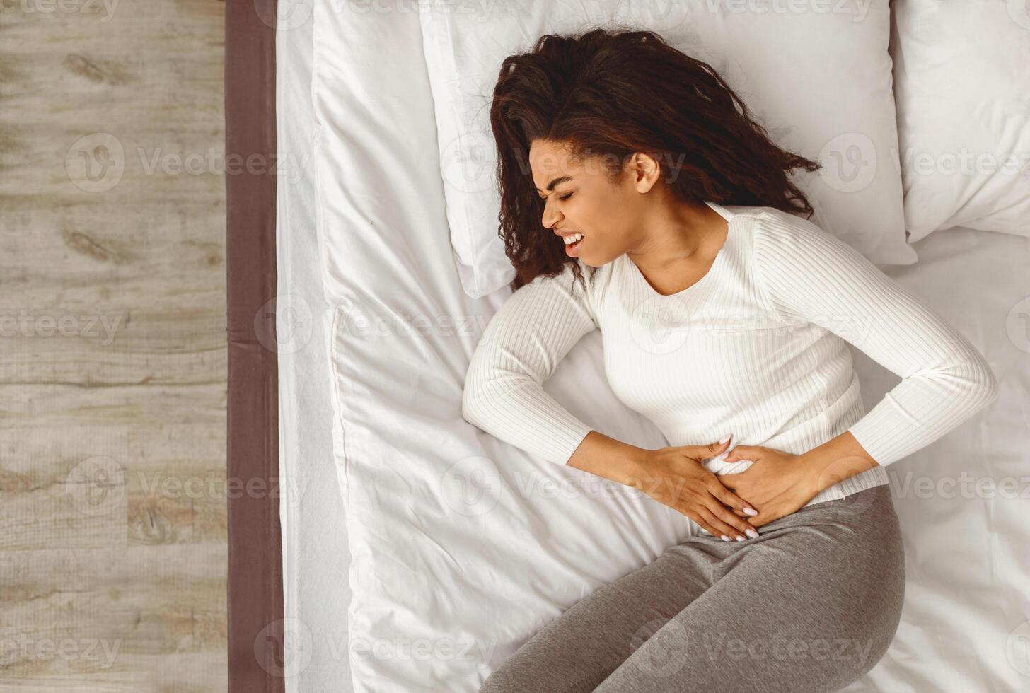 Above top high angle view of upset black woman suffering from strong abdominal pain, touching her tummy, lying on back in bed. Sad lady feeling acute stomachache, free copy space photo