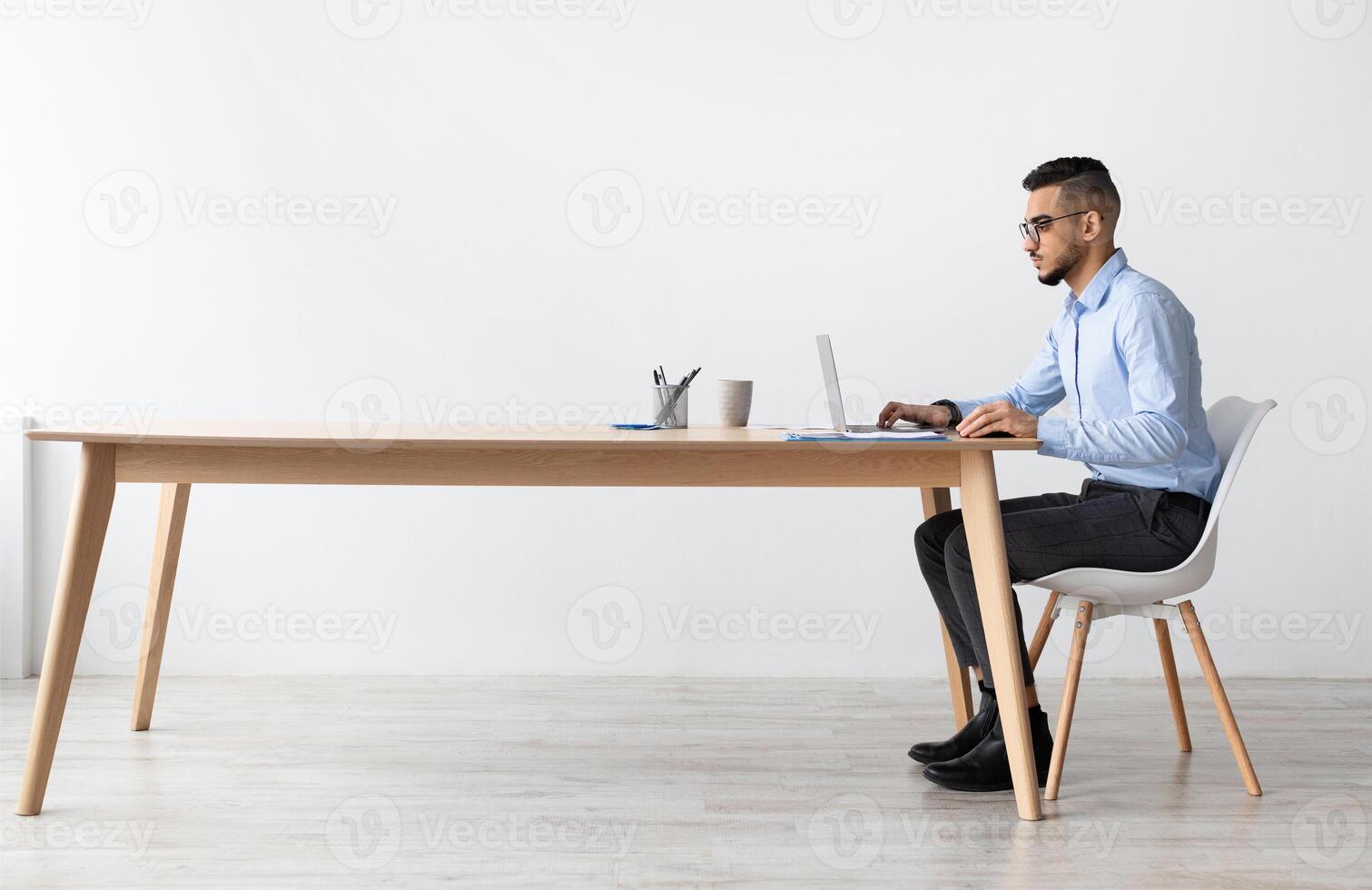 Profile Side View Portrait Of Focused Middle Eastern Male Manager In Glasses Working On Laptop Computer In Modern Office, Serious Guy Sitting At Desk And Using Pc, Looking At Screen Watching Webinar photo