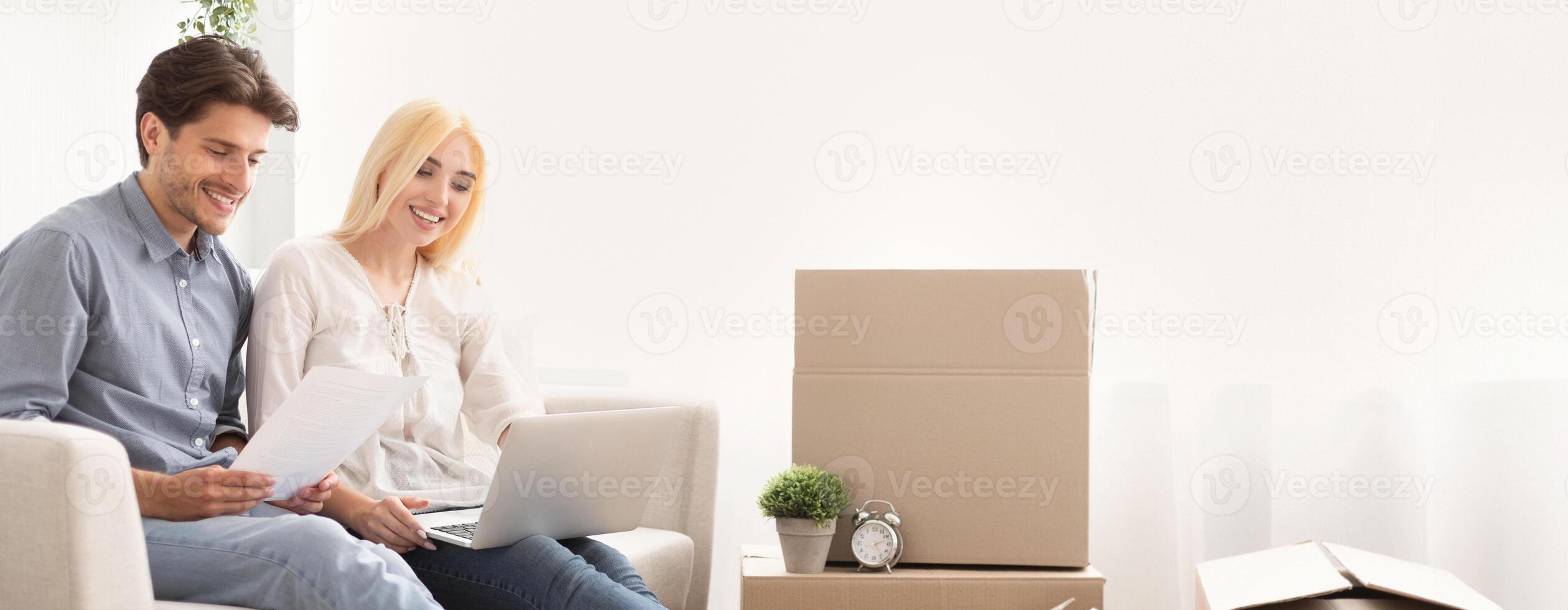 A young couple smiles while sitting on a couch, looking at documents and using a laptop. In the background, there are unpacked cardboard boxes and a small plant, indicating a recent move. photo