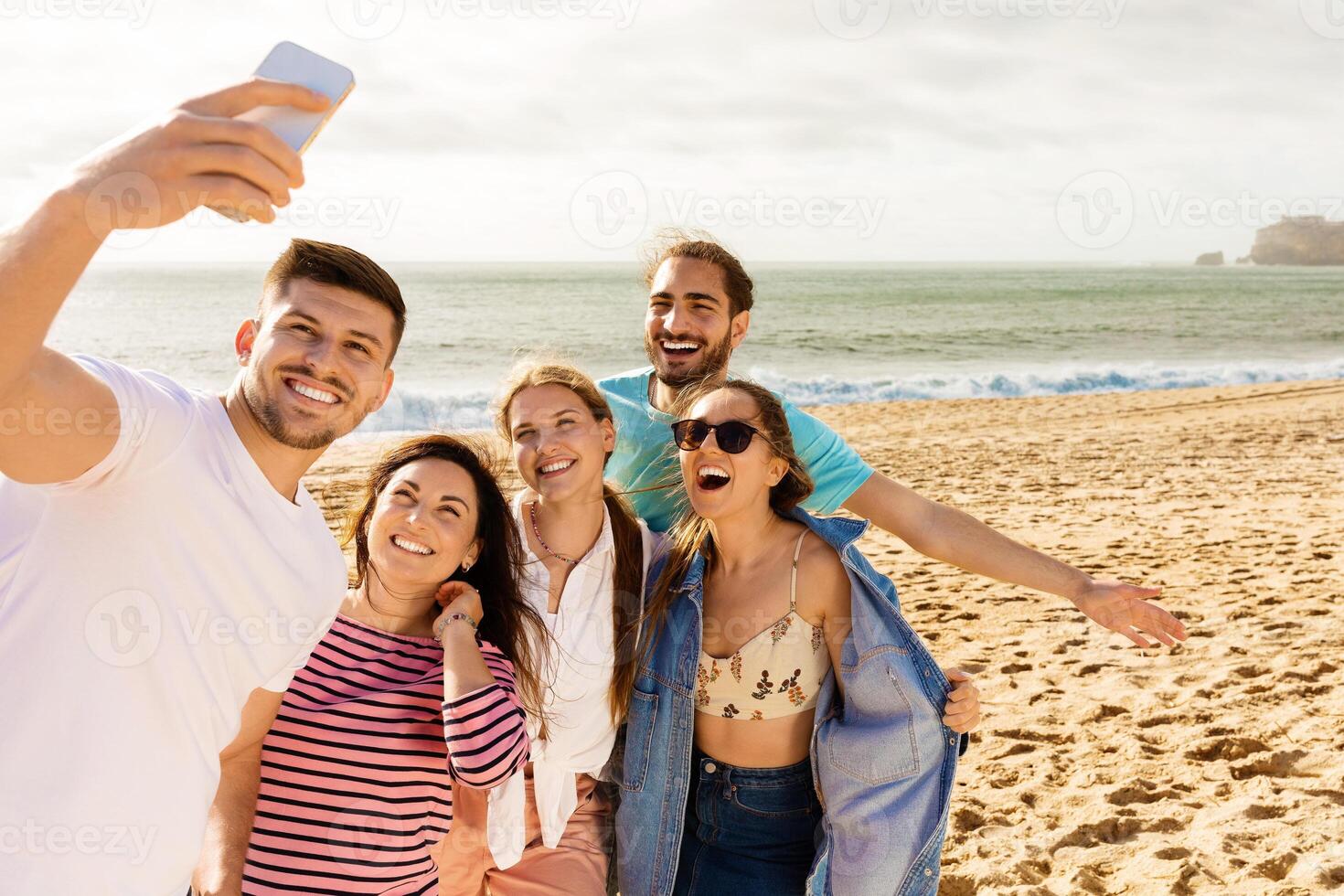 A group of five friends gathers on the sandy beach, smiling and taking selfies as the sun sets behind them, creating a joyful and lively atmosphere during their seaside outing. photo