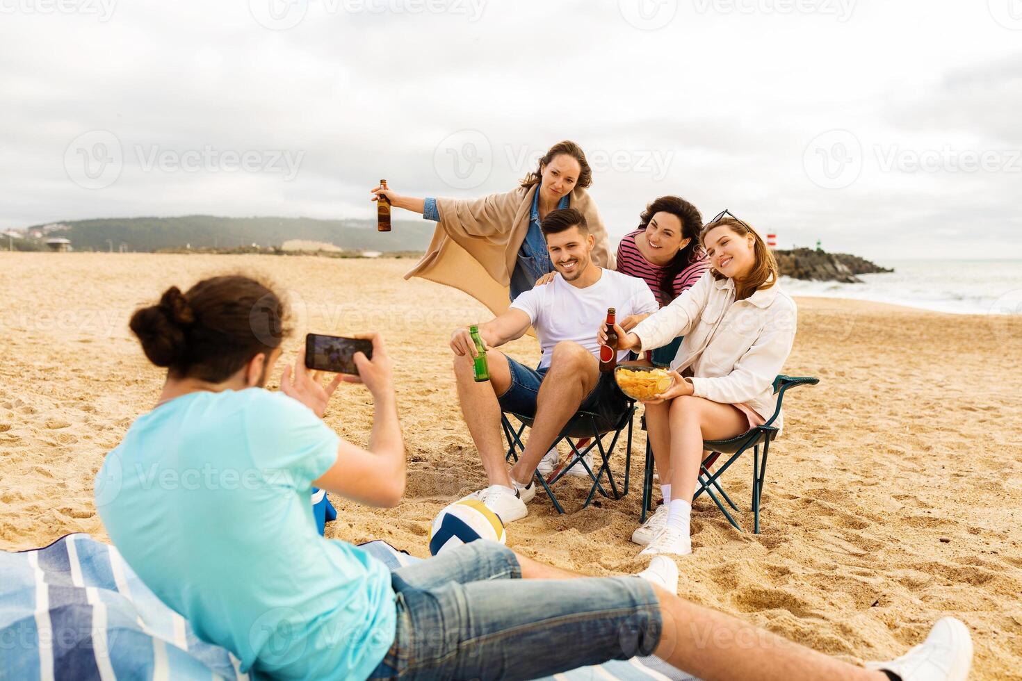A group of friends sits on the sand, smiling and posing for a picture. They have snacks and drinks as they relax and enjoy their time together on a cloudy summer day. photo
