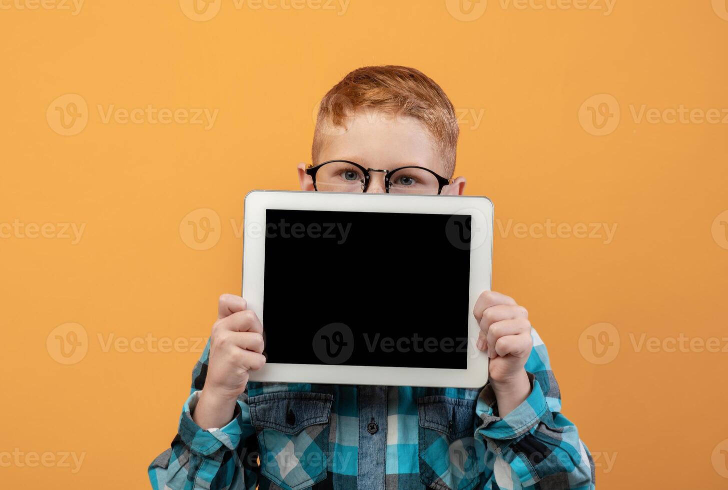 Ginger boy in eyeglasses hiding behind digital tablet with empty screen over yellow studio background. Cute redhead schoolboy showing pad, mockup, advertisement for cute application photo