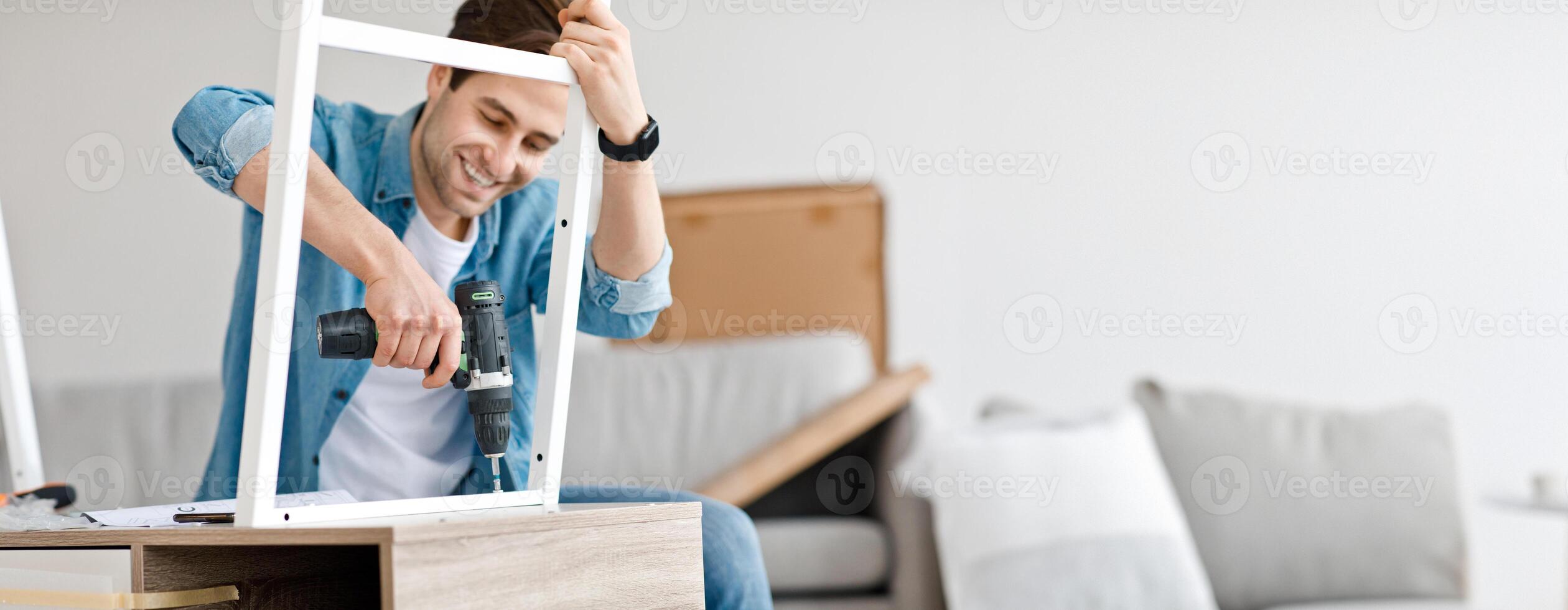 Assembling chipboard furniture using cordless screwdriver at home. Smiling young busy man with smart watch screwing details of table with electric drill, in interior of living room, copy space photo