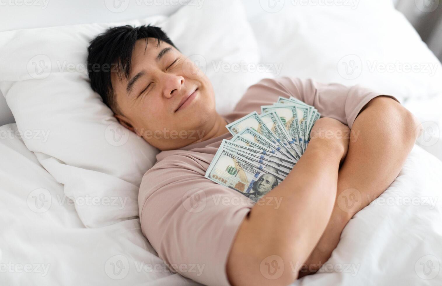 Happy sleeping middle aged chinese man hugging bunch of cash dollars, lying on bed, asian male keeps money safe, top view. Savings, financial literacy, economy concept photo