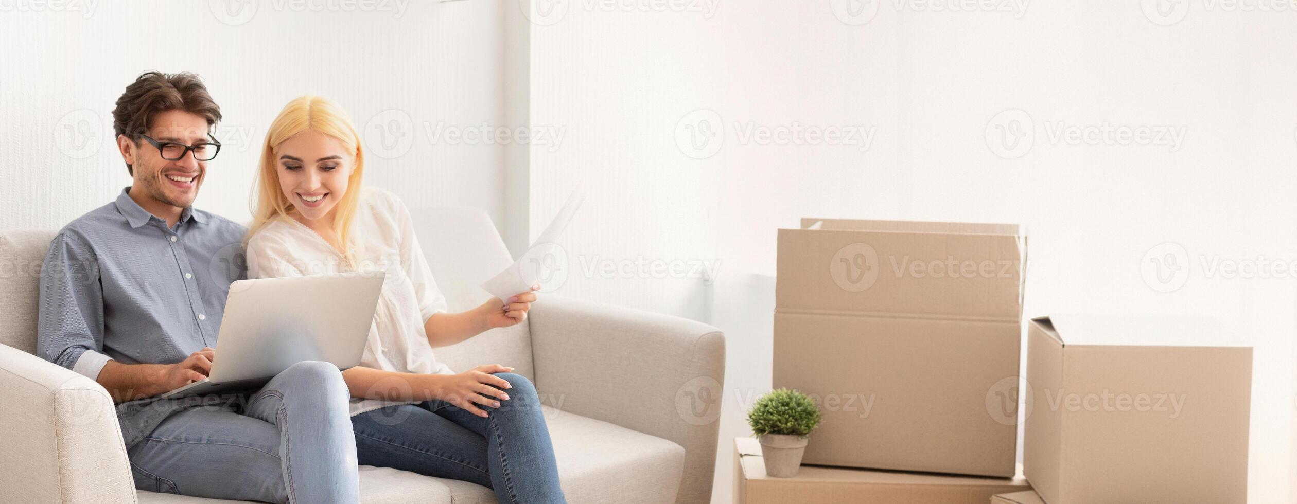 Cheerful Husband And Wife Browsing On Laptop Sitting On Couch In New Flat. Copy Space photo