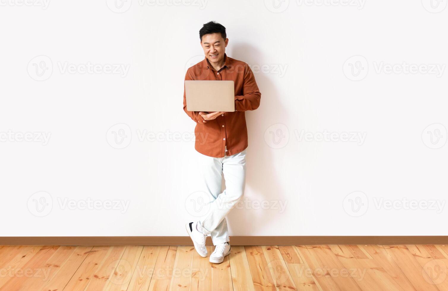 Full length shot of cheerful asian man in casual wear using laptop computer for online work or communication, male standing against white studio wall, copy space photo