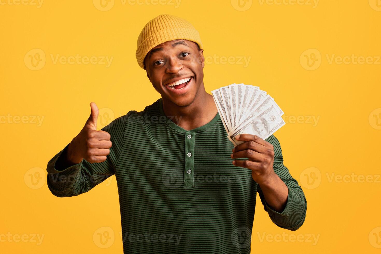 Big Profit. Cheerful Young Black Man Holding Dollar Cash Money And Showing Thumb Up, Excited African American Millennial Guy Celebrating Financial Success, Standing Over Yellow Studio Background photo