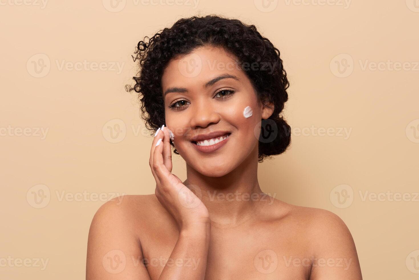 A young woman with curly hair smiles while applying moisturizer to her face. She stands against a soft beige background, showcasing her natural beauty and skincare routine. photo
