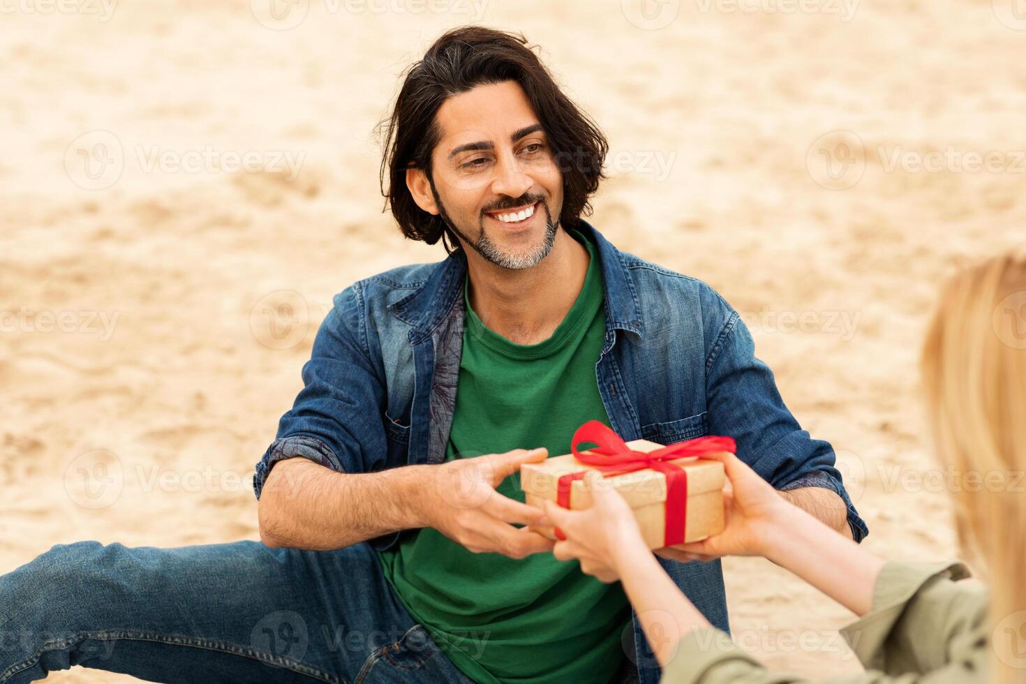 A man sitting on a sandy beach smiles as he receives a beautifully wrapped gift with a ribbon from a woman. The warm sun shines down on them, creating a joyful atmosphere. photo