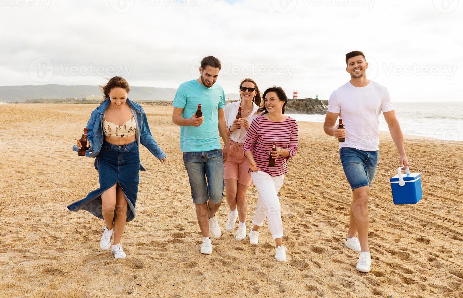 A group of five friends strolls along a sandy beach, laughing and carrying drinks. The sun shines brightly as they enjoy a warm day by the coast, sharing good times together. photo