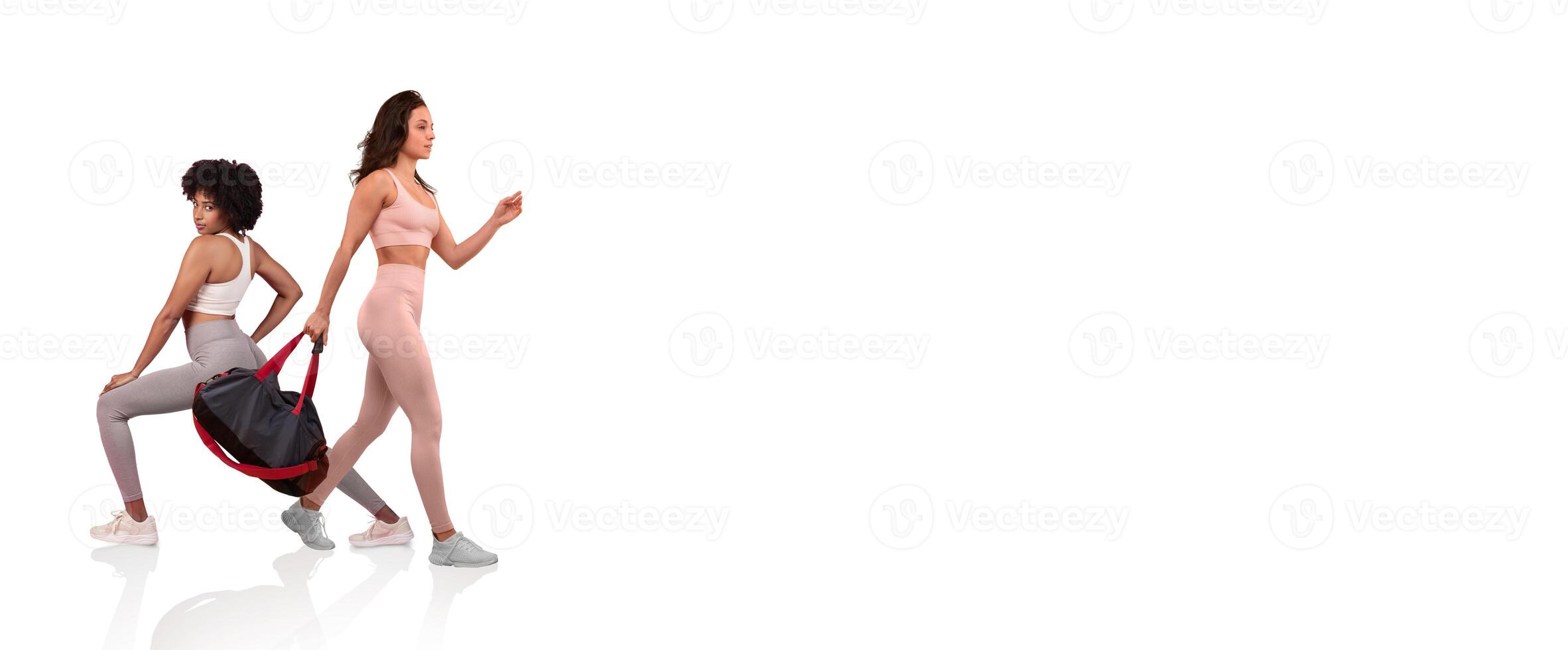 Two women engage in a workout session, one performing a lunging exercise while the other walks confidently with a gym bag. They are dressed in matching activewear in a bright environment. photo