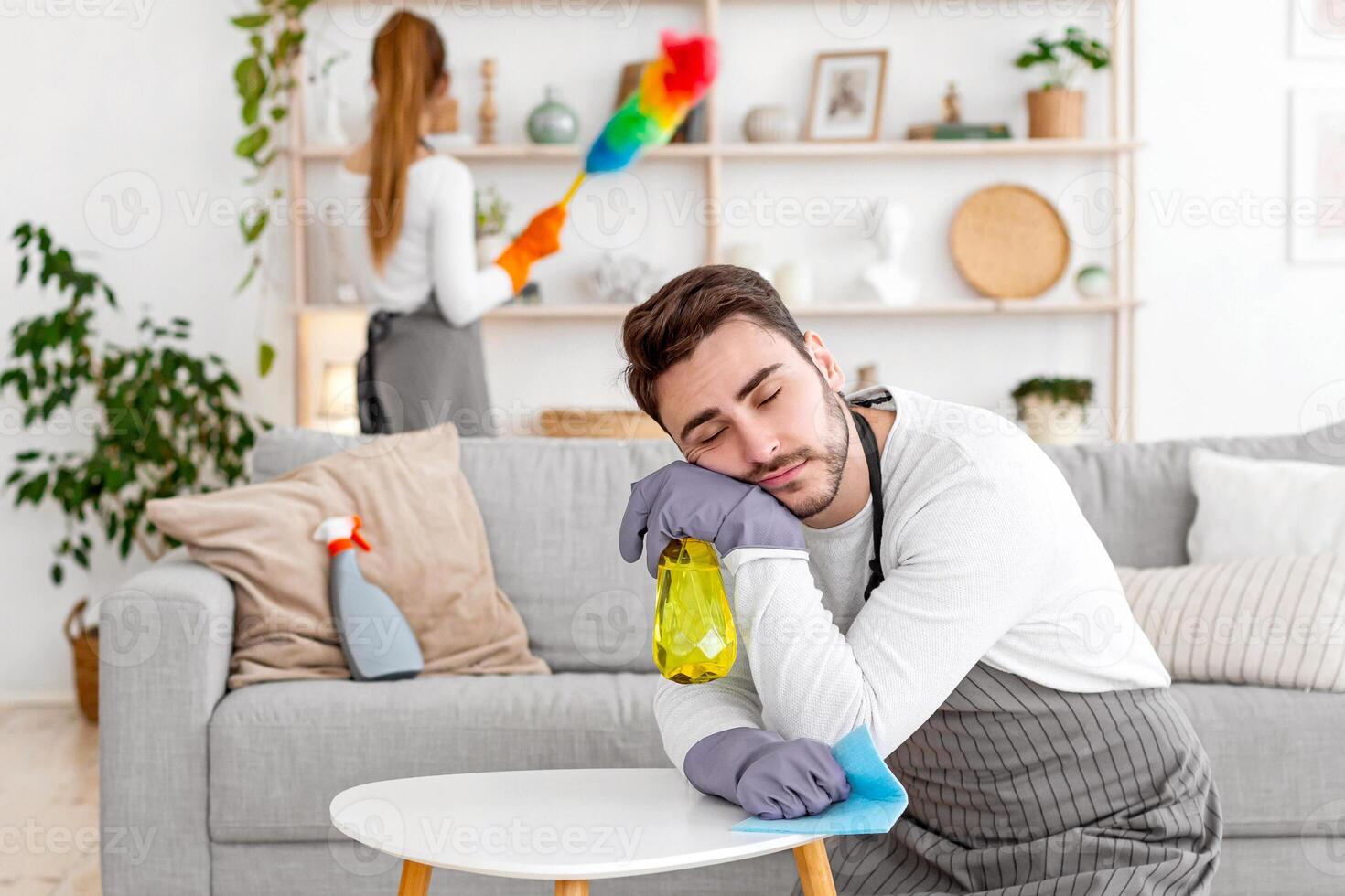 Household chores and tired. Man in apron and gloves sleeping on his knees and table, wife wipes furniture in living room, free space photo