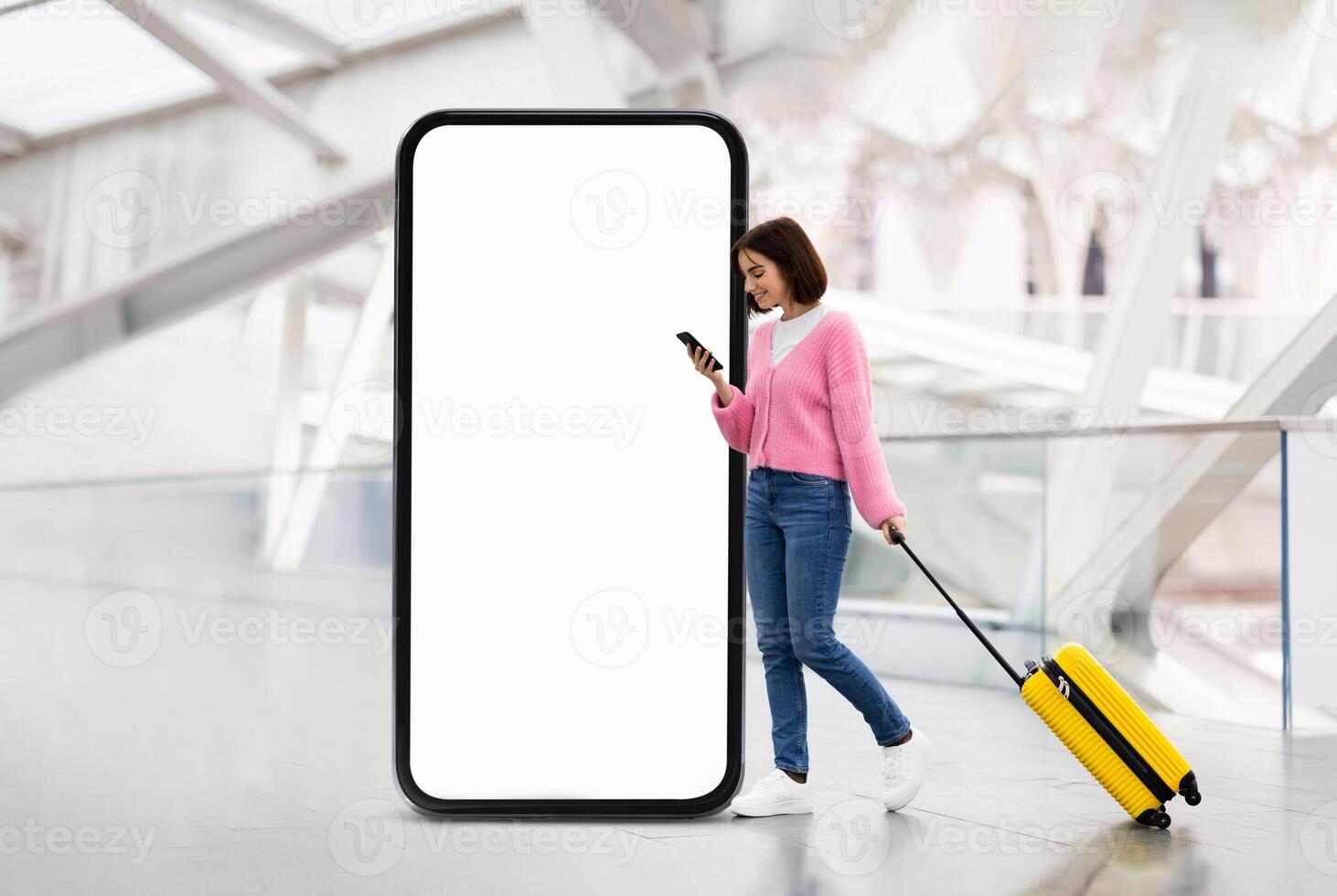 A young woman standing indoors at a modern location is using her phone while pulling a yellow suitcase. She is located next to a large blank digital advertising screen, suggesting a travel context. photo