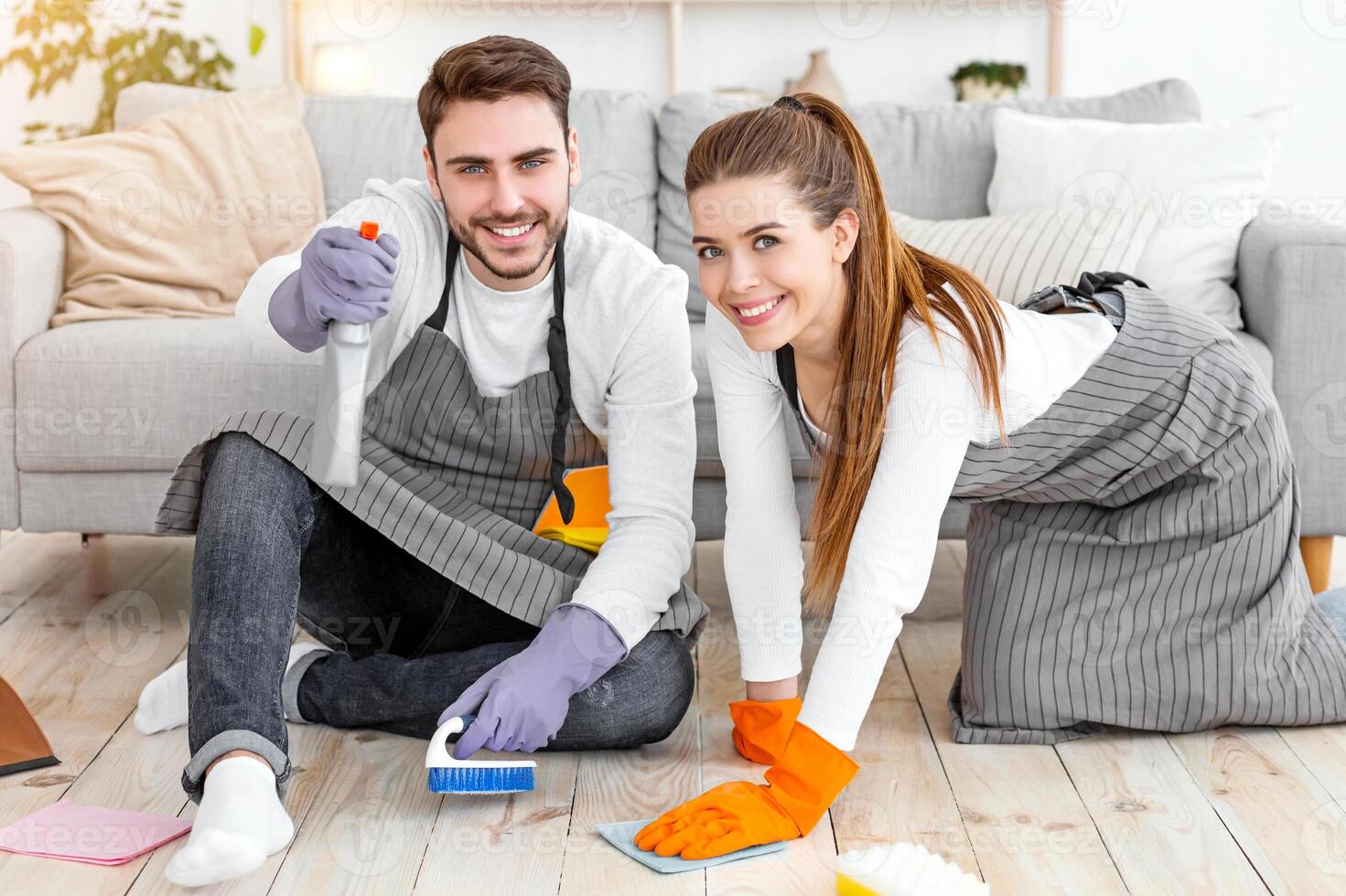 Easy cleaning with pleasure. Happy man and woman in aprons and rubber gloves wash floor with cleaning supplies in living room interior, free space photo