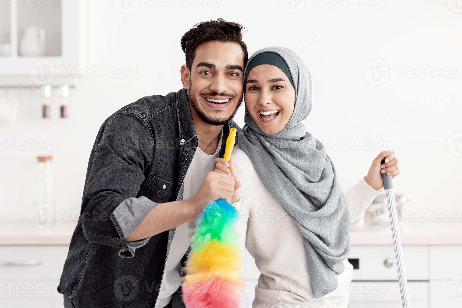 Happy arab lovers enjoying house-keeping at their new home, cheerful lady in hijab and handsome bearded guy holding mop and dust brush as microphones, singing while cleaning kitchen, copy space photo