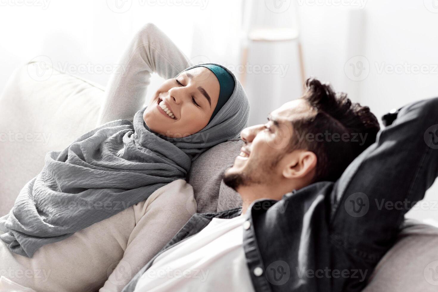 Relaxed muslim family resting after moving to their new house, sitting on new couch among paper boxes with stuff and having break while unpacking belongings, leaning back and smiling, copy space photo