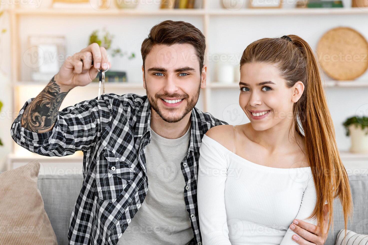 Buying or renting real estate. Happy millennial husband and wife sitting on sofa and looks at camera, man showing keys to new apartment in interior of modern living room, free space photo