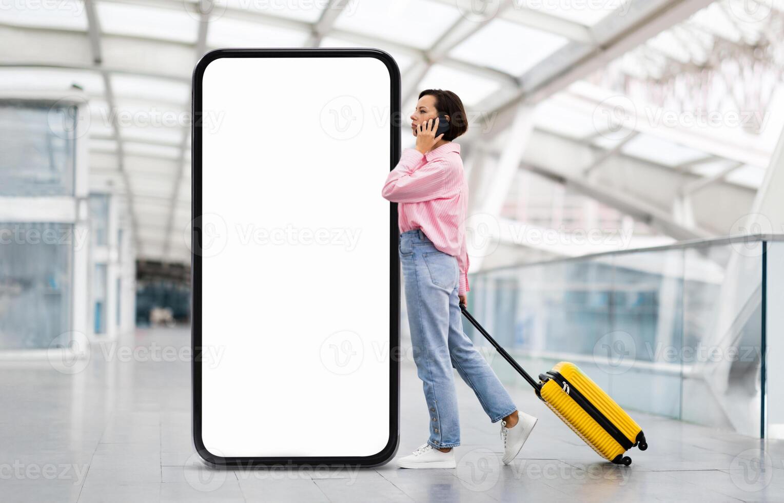 A traveler stands at the airport casually talking on a smartphone, pulling a yellow suitcase, passing by huge phone with blank screen photo