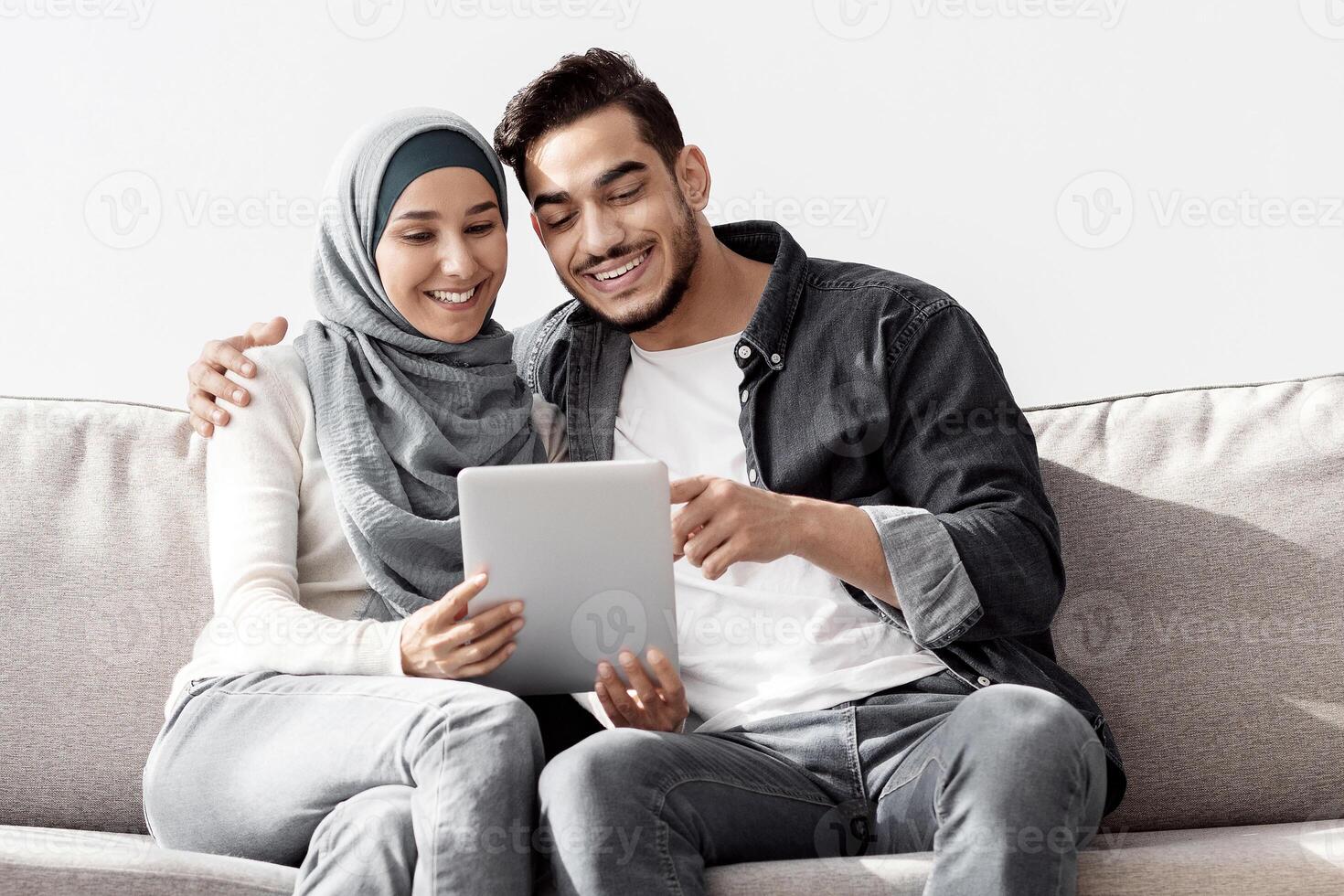 Joyful muslim family sitting on couch in new apartment, using pad, copy space. Happy middle-eastern young man and woman in hijab buying furniture for house, unpacking stuff after moving photo