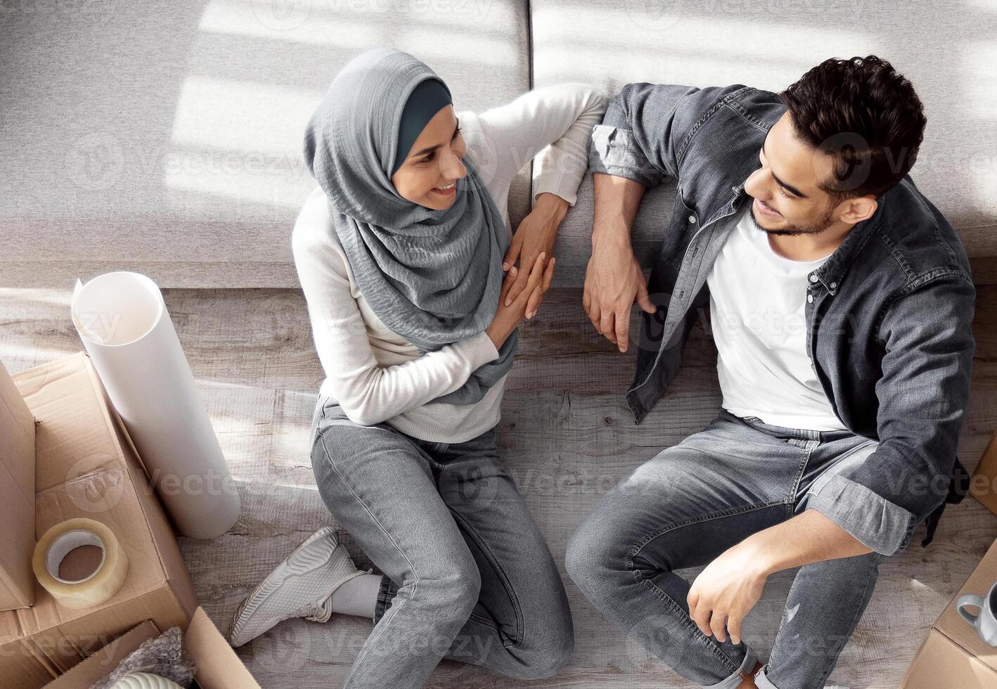 Top view of young muslim family sitting on floor by couch among moving supplies, having break while moving to their new apartment. Happy arab man and woman in hijab with paper boxes photo