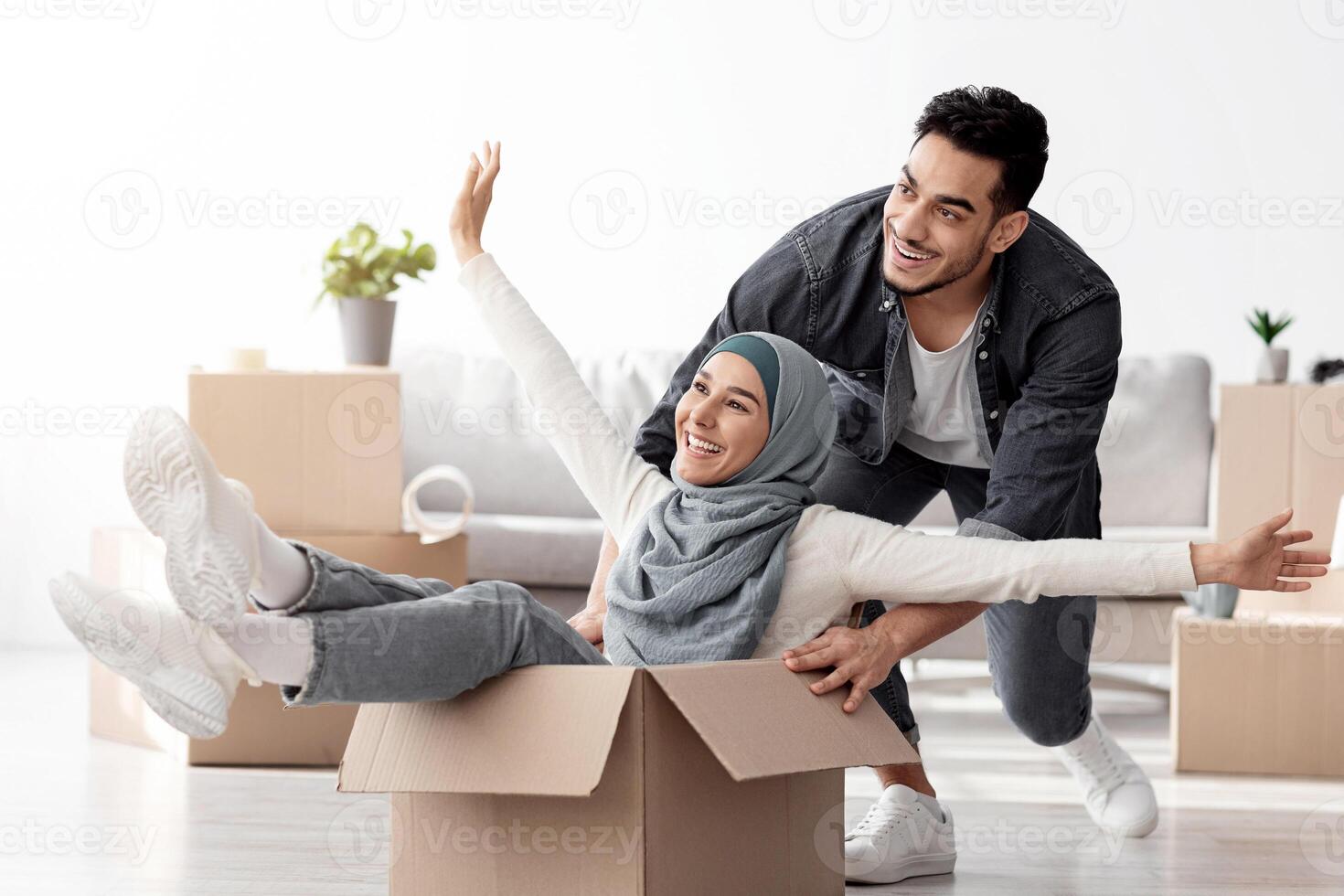 Emotional middle-eastern young family having fun while unpacking stuff in their new house. Happy arab guy and excited lady in hijab sitting in paper box, enjoying moving. Apartment, house, real estate photo