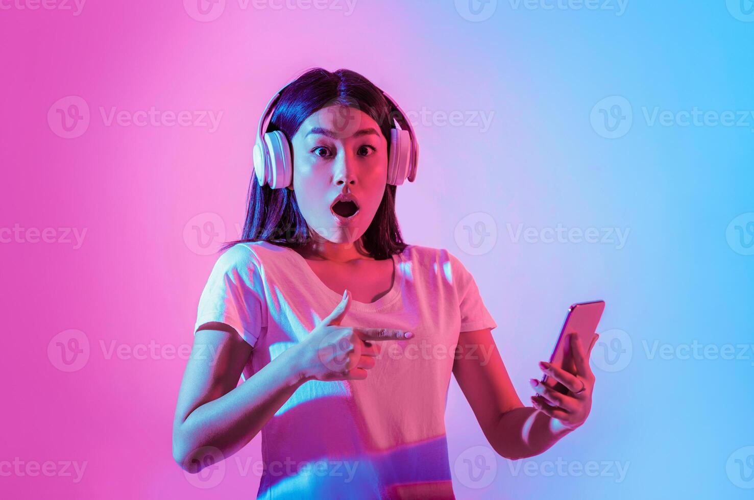 Facial expression. Surprised asian lady with open mouth in large wireless headphones, points finger in smartphone in neon, studio shot photo