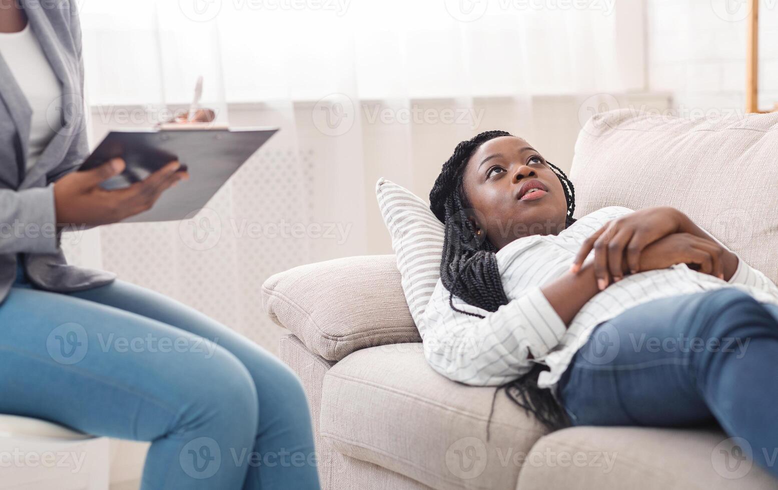 At the psychologist. Depressed young afro woman lying on couch during psychotherapy session while doctor taking notes, free space photo