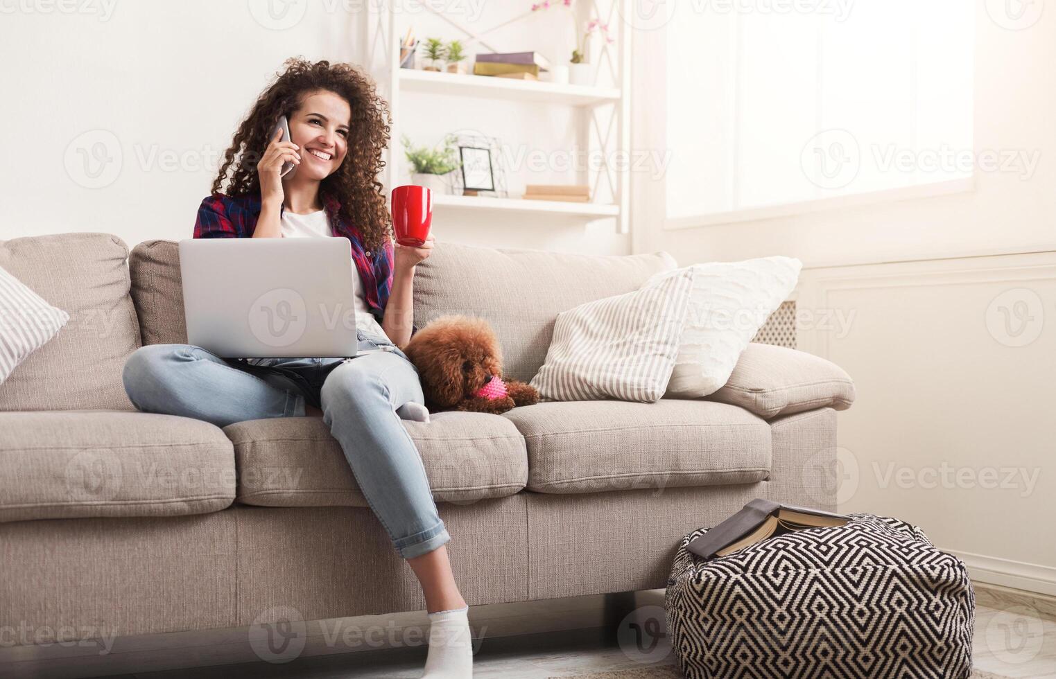 Happy girl with smartphone, laptop and dog at home. Curly woman talking on mobile, surfing the web on couch with her puppy, copy space photo