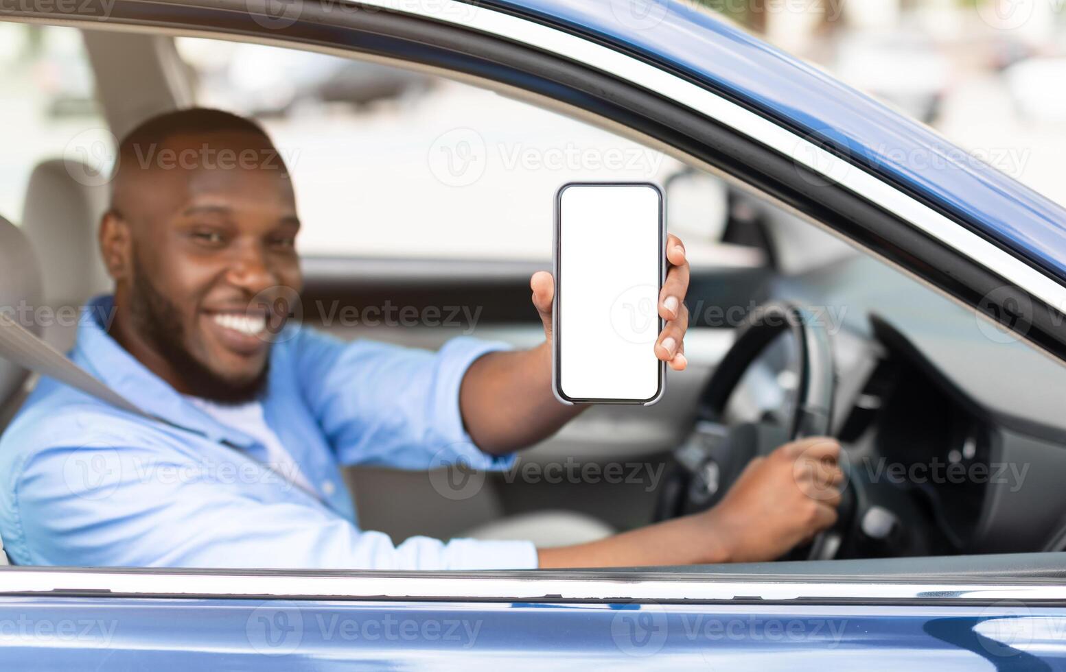 Closeup of young smiling African American man showing mobile phone with white empty screen for mock up sitting on driver seat in new luxury car, holding gadget in hand, selective focus on device photo
