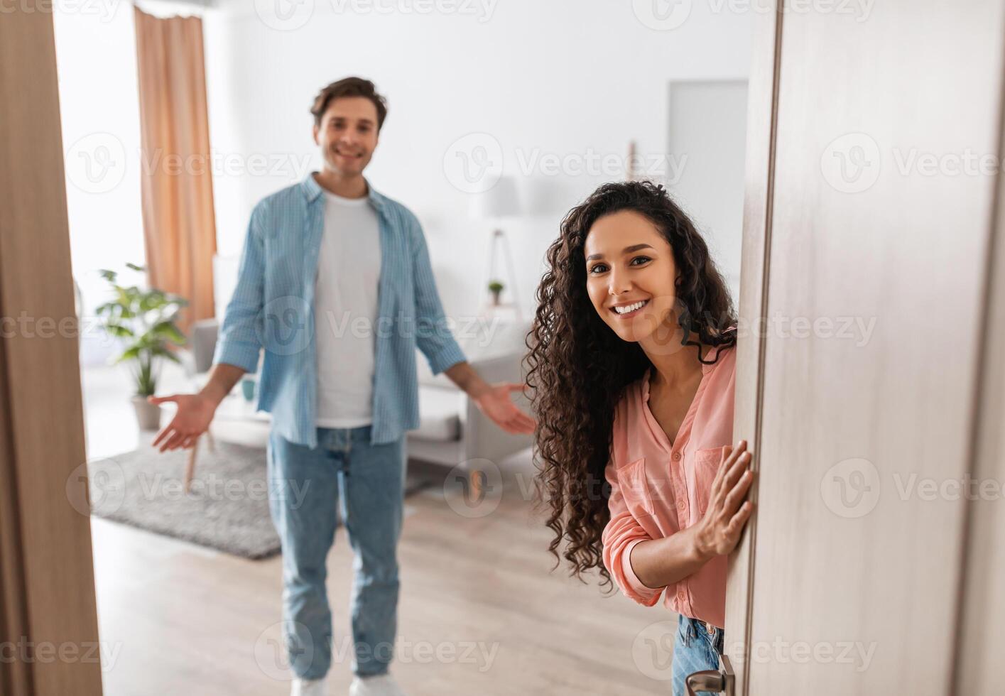 Portrait of cheerful couple inviting guests to enter home, happy people standing in doorway of modern flat, looking out waiting for visitor to come in, receiving friend, lady peeking out of front door photo