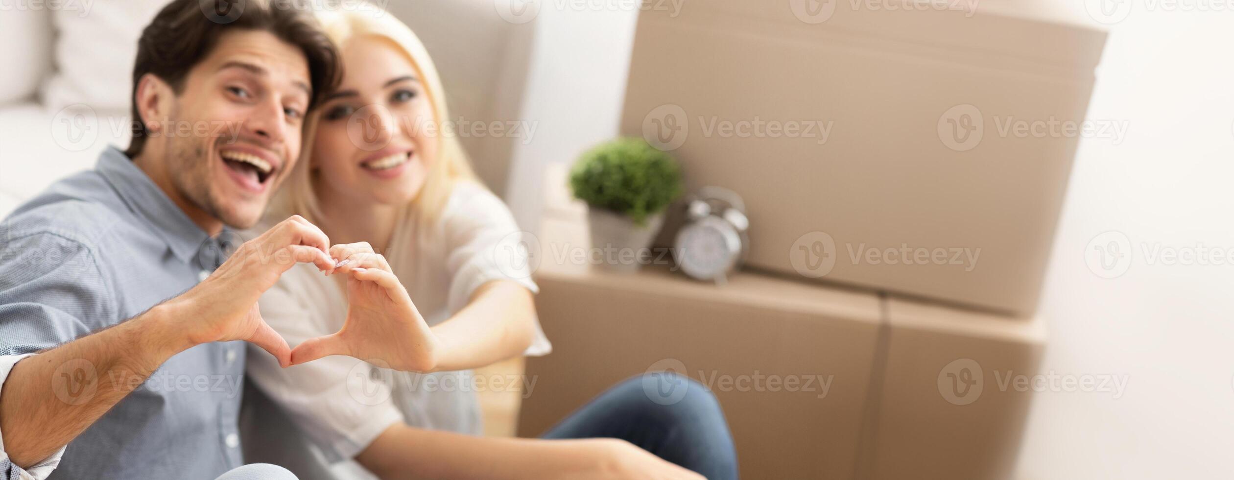 Loving Husband And Wife Gesturing Heart Shape With Hands Sitting Among Moving Boxes In New Home. House Owners Concept, Copy Space photo