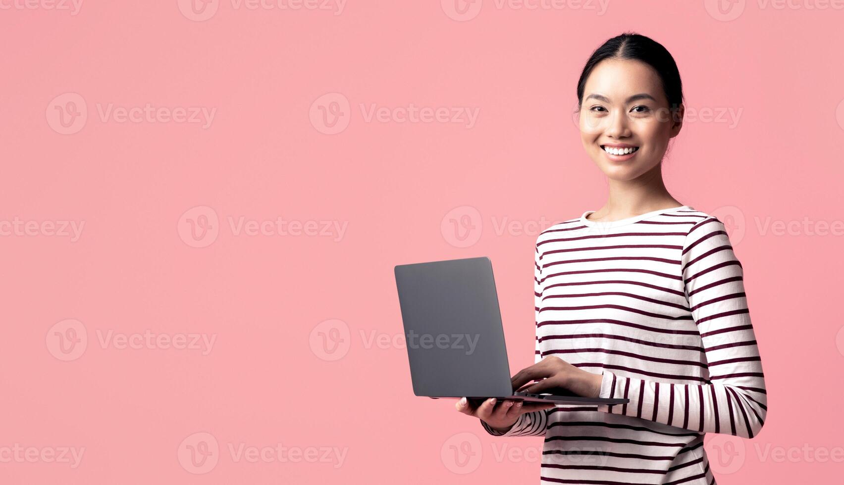 Portrait Of Young Smiling Asian Woman With Laptop Computer In Hands Standing Isolated Over Pink Background, Korean Female Working Online On Computer Or Browsing New Website, Panorama With Copy Space photo