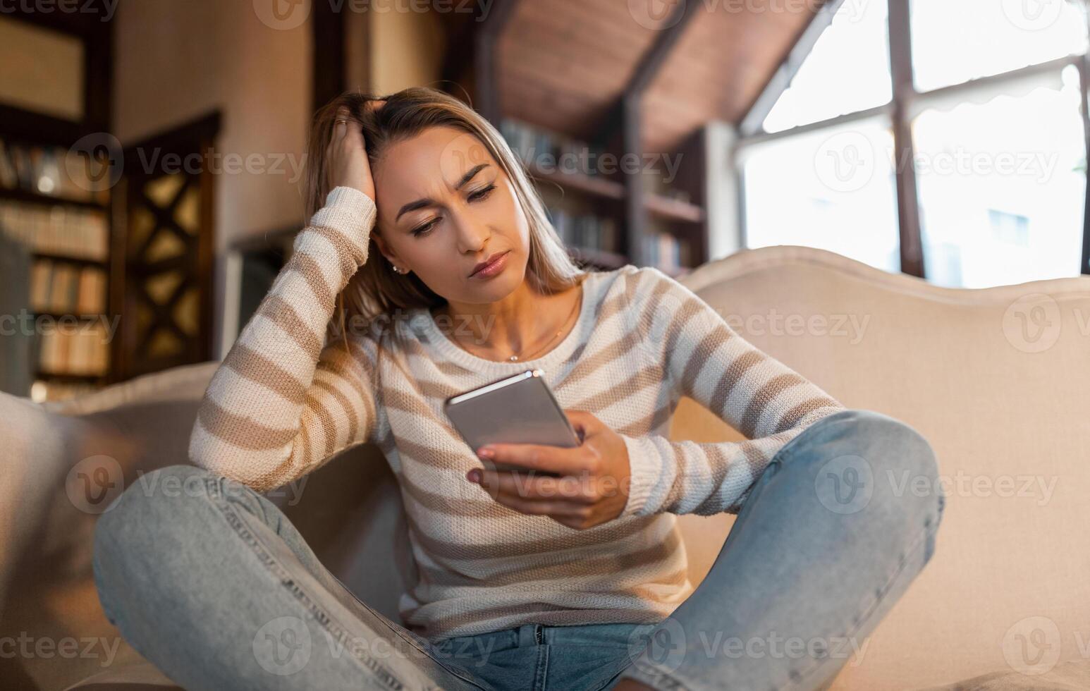 Bad News Concept. Upset Confused Woman Holding Smartphone, Looking At Mobile Phone Screen With Worried Expression, Touching Head, Sad Adult Female Reading Unplesant Message Sitting On Couch photo
