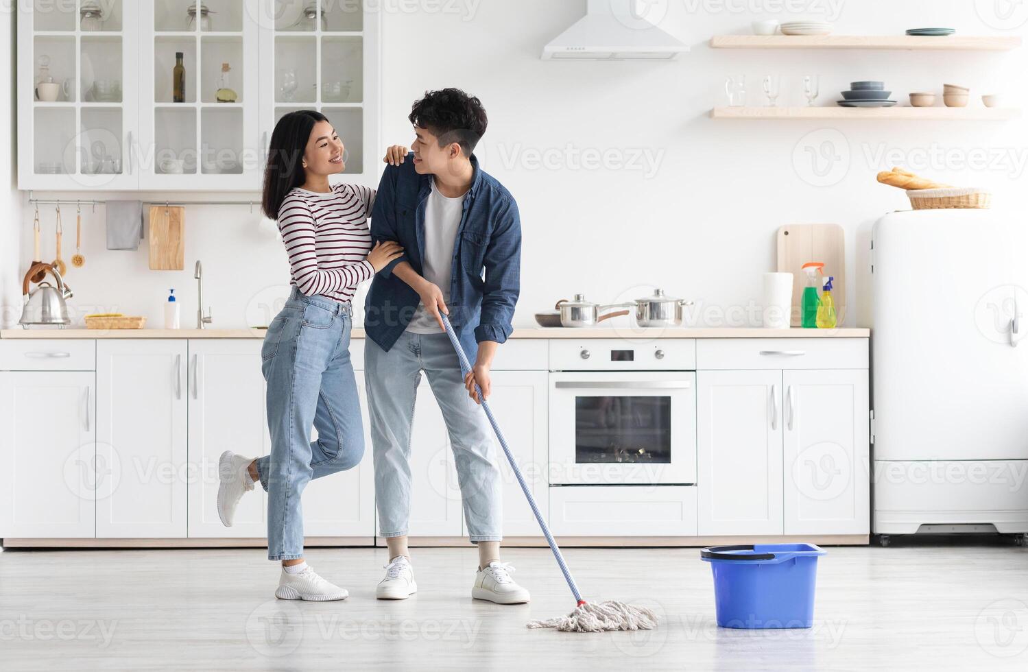 Beautiful loving asian young couple mopping floor in kitchen together, full length photo, copy space. Romantic cheerful chinese lady hugging her smiling boyfriend while cleaning house photo