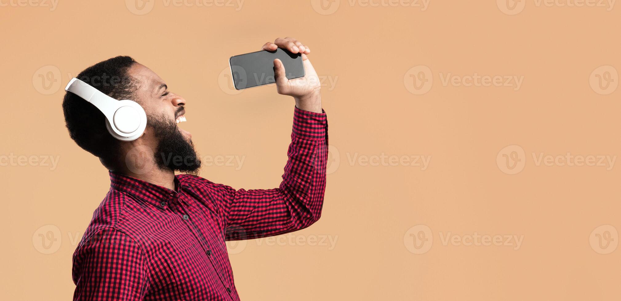 Excited emotional black man singing his favorite song, using smartphone as microphone, yellow studio background photo