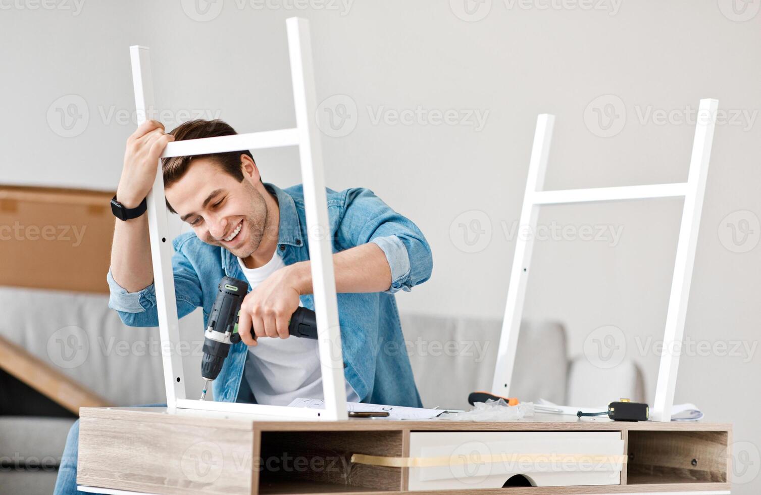 Manual job at home, handyman, carpenter and good service. Smiling millennial handsome man with smart watch assembles furniture with electric screwdriver, work with pleasure in living room, empty space photo