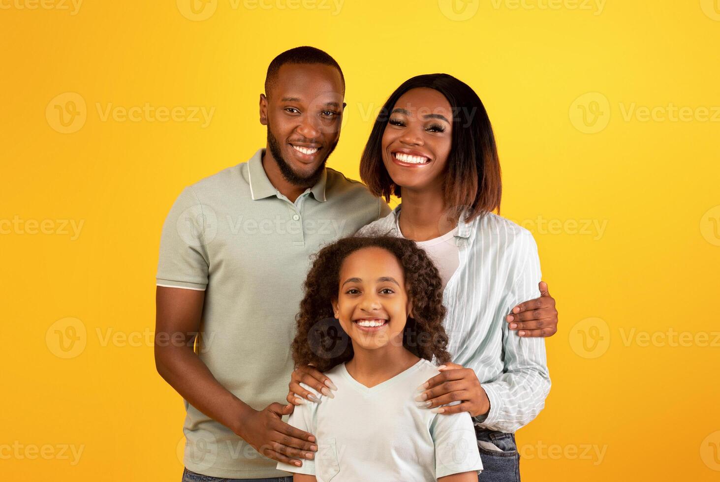 Family love concept. Portrait of black family with daughter hugging and smiling at camera standing isolated on yellow background. Mum and dad posing with child in studio photo