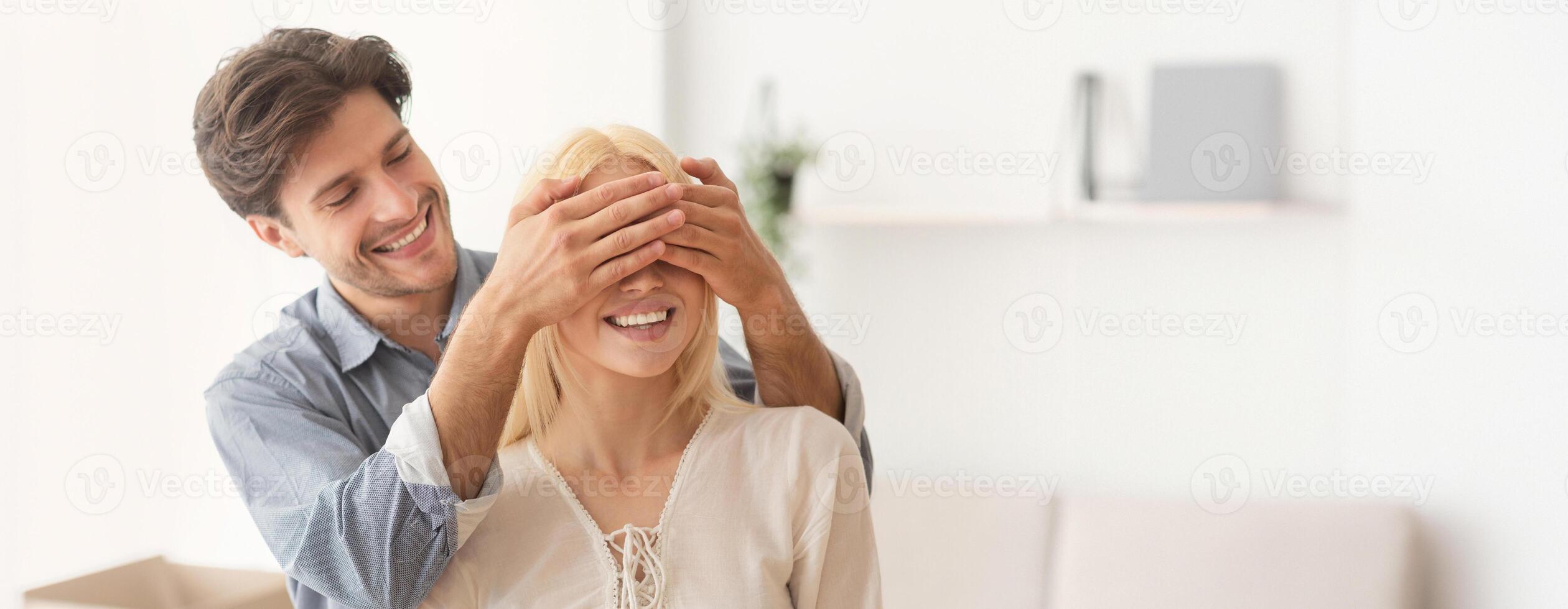 A cheerful man covers a woman eyes from behind, creating a delightful surprise. They are in a modern, well-lit room, sharing a joyful moment together, filled with laughter and excitement, copy space photo