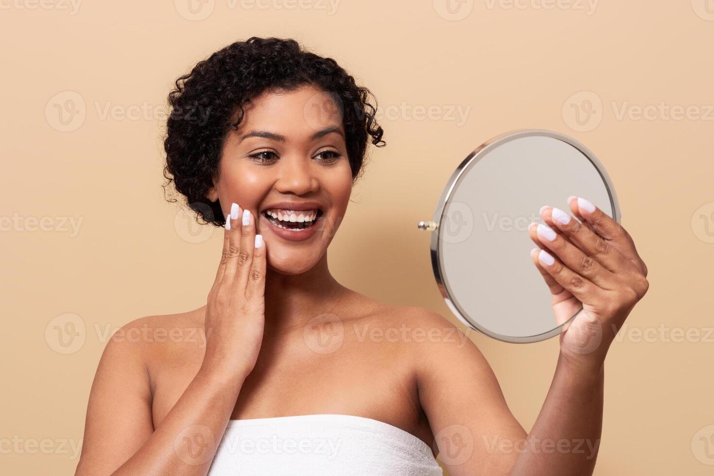 A woman with curly hair admires her reflection in a hand mirror while applying skin care. She has a joyful expression as she enhances her natural beauty, enjoying her routine. photo