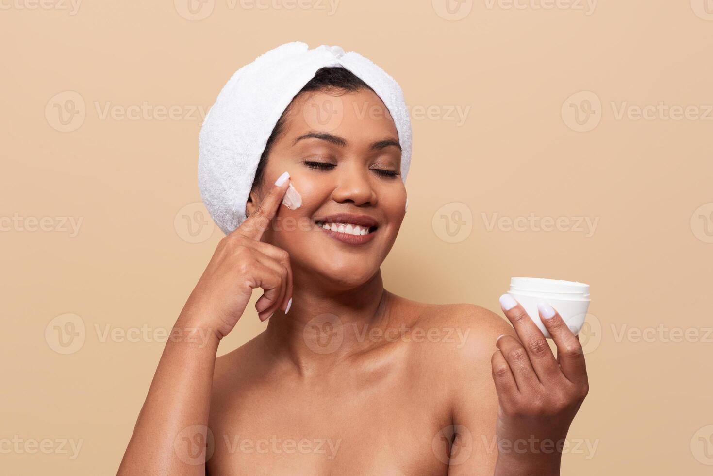 A woman with a towel wrap on her head is applying moisturizer to her face while smiling. She holds a jar of cream and enjoys her skincare routine in a warm-toned setting. photo