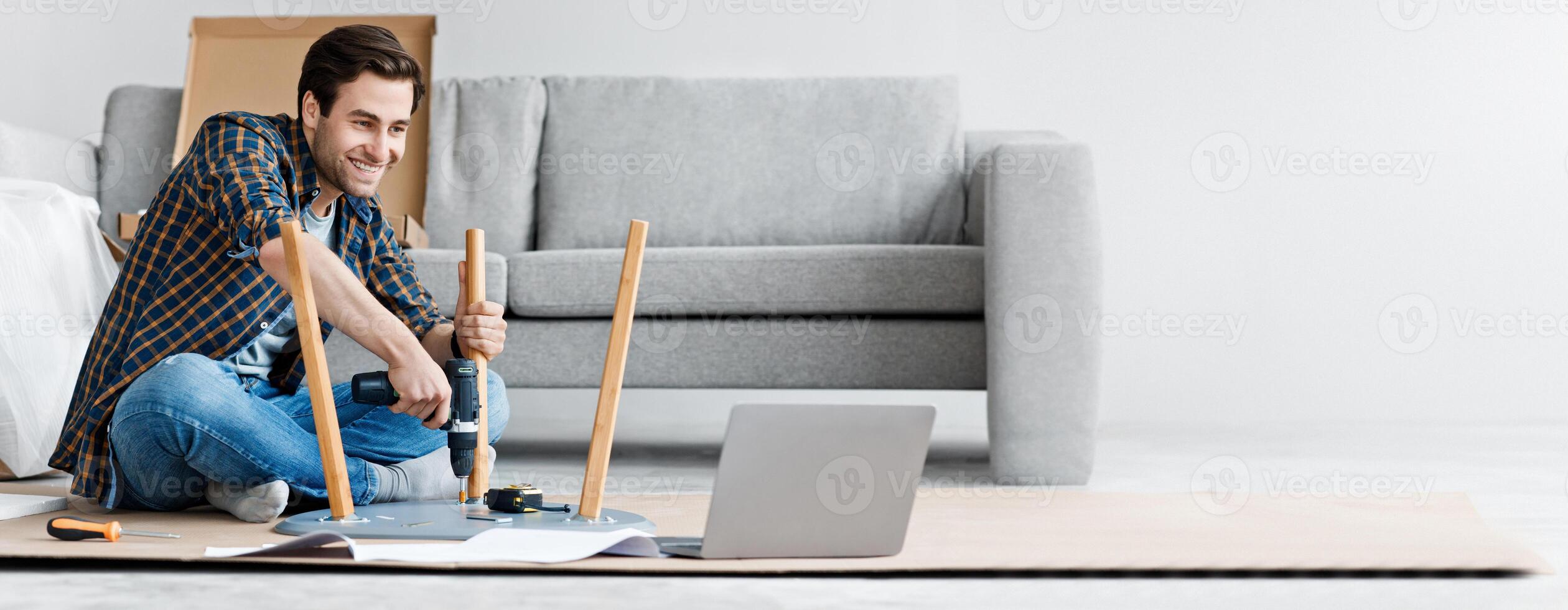 Assemble furniture, collects chair, handyman with tools repairs stool. Happy young guy screwing detail with electric drill on floor in living room and looking at laptop, watch instruction, copy space photo