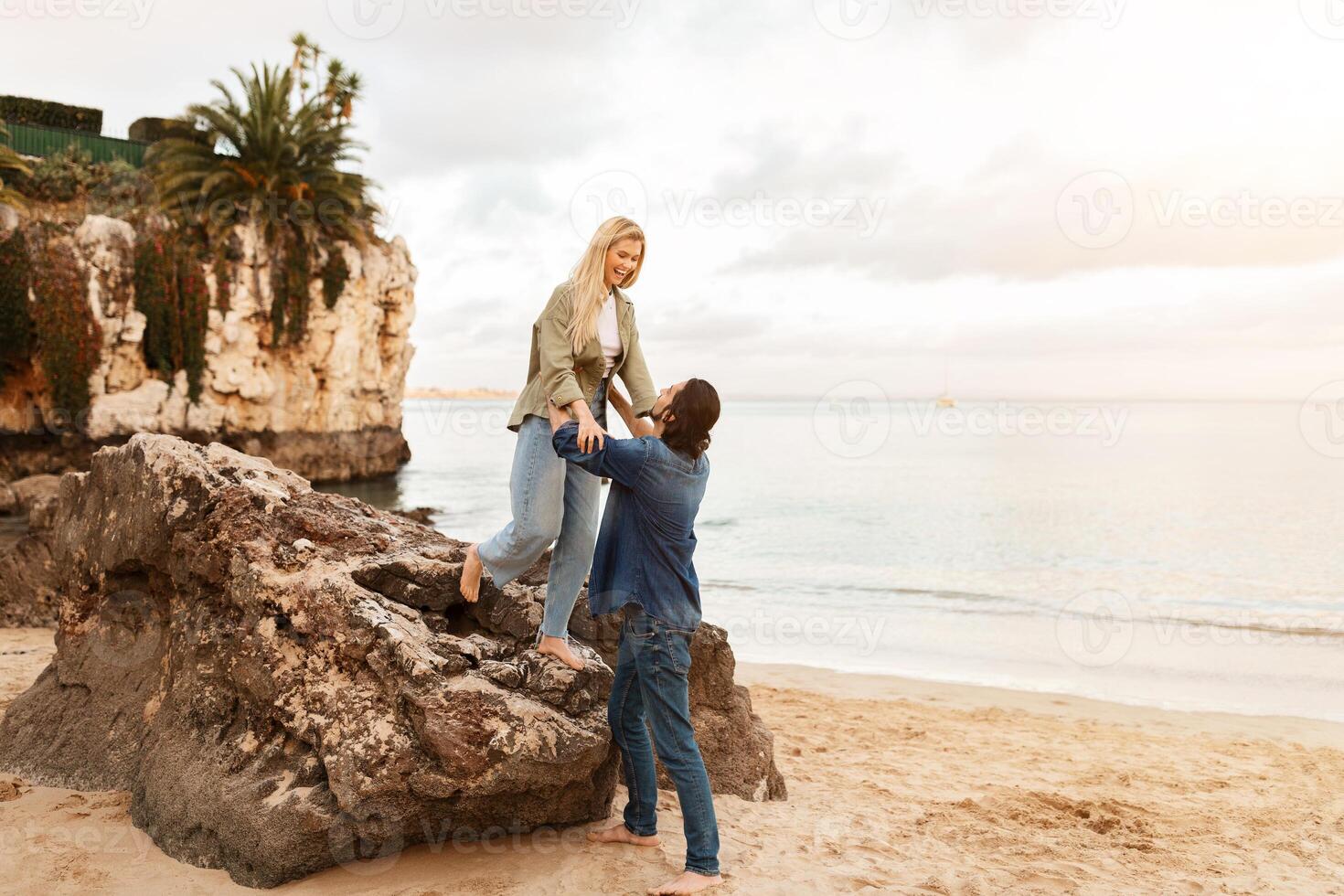 A couple shares a joyful moment on the beach as the sun sets. The woman stands on a rock while the man helps her down, surrounded by gentle waves and a serene atmosphere. photo