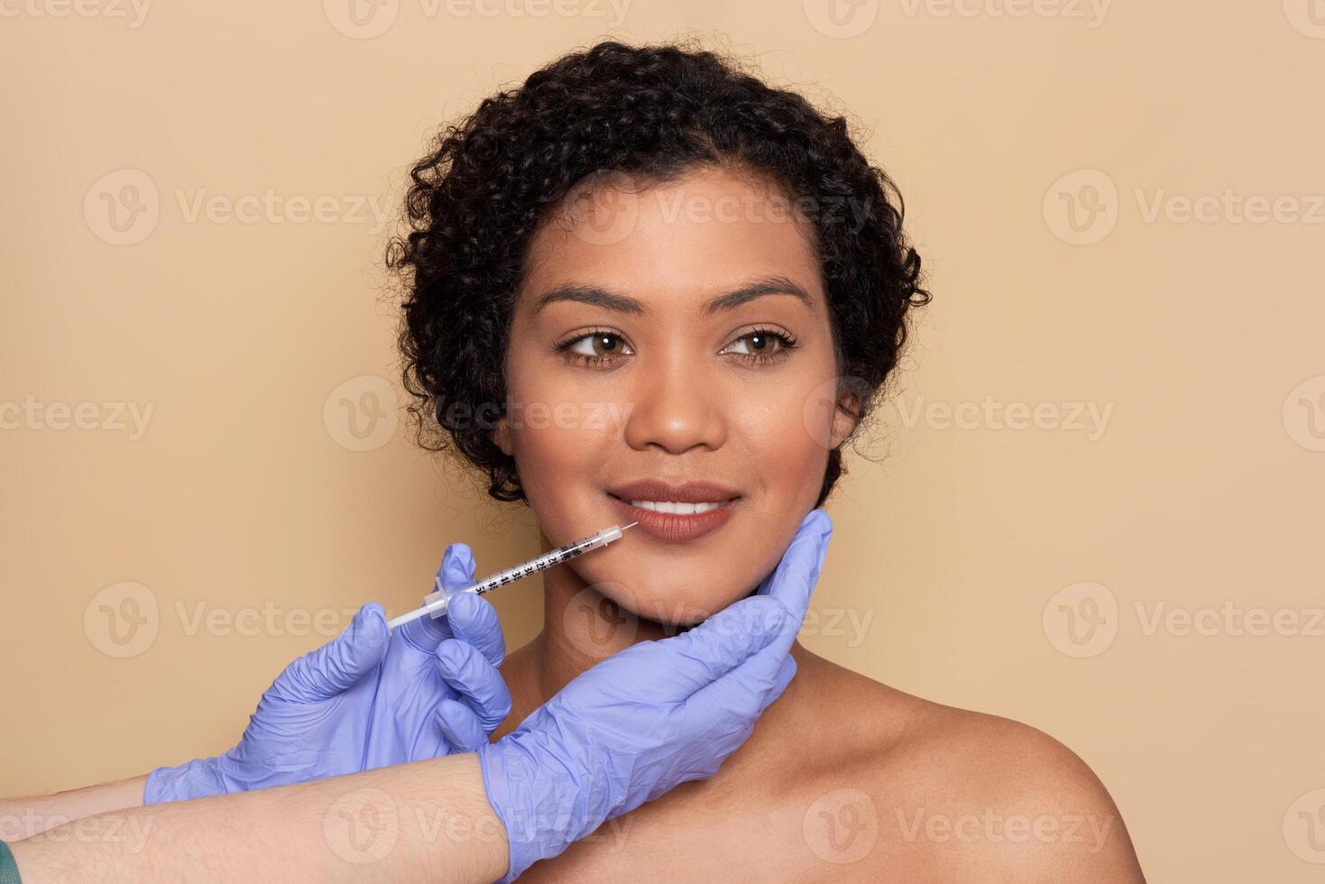 A woman with curly hair is receiving a cosmetic procedure at a clinic. A professional with gloved hands is administering an aesthetic treatment to enhance her facial features. photo