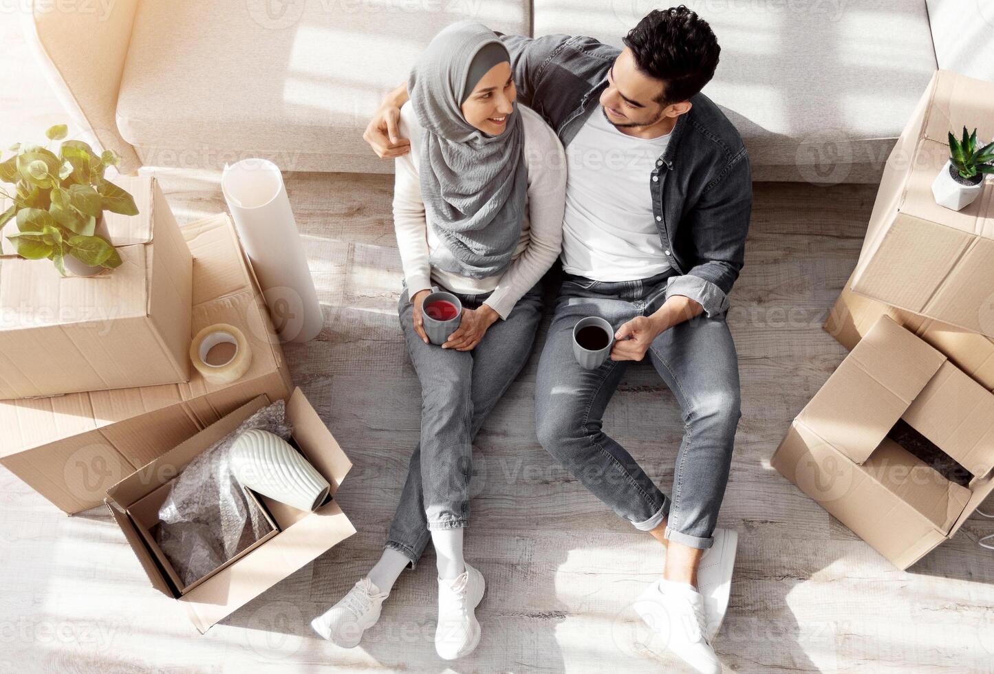 Top view of loving arab husband and wife in hijab sitting on floor by couch in living room, having break while unpacking stuff at new house, hugging and drinking coffee. Moving, recolating concept photo
