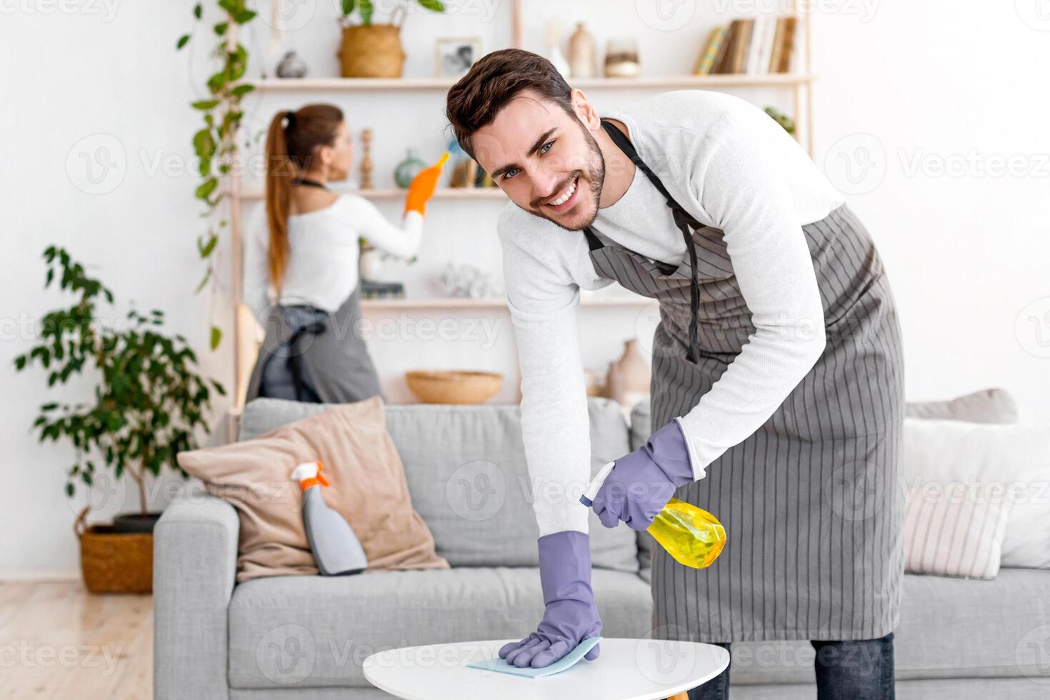 Stay at home and cleaning flat spending quarantine together. Cheerful man in apron and rubber gloves with spray and rag wipes dust with wife, near cleaning supplies in interior of living room photo