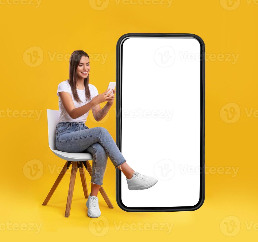 Cheerful smiling female sitting on chair near big cell phone with empty white screen, using mobile device, checking new cool app on orange studio background, mock up for website or application design photo