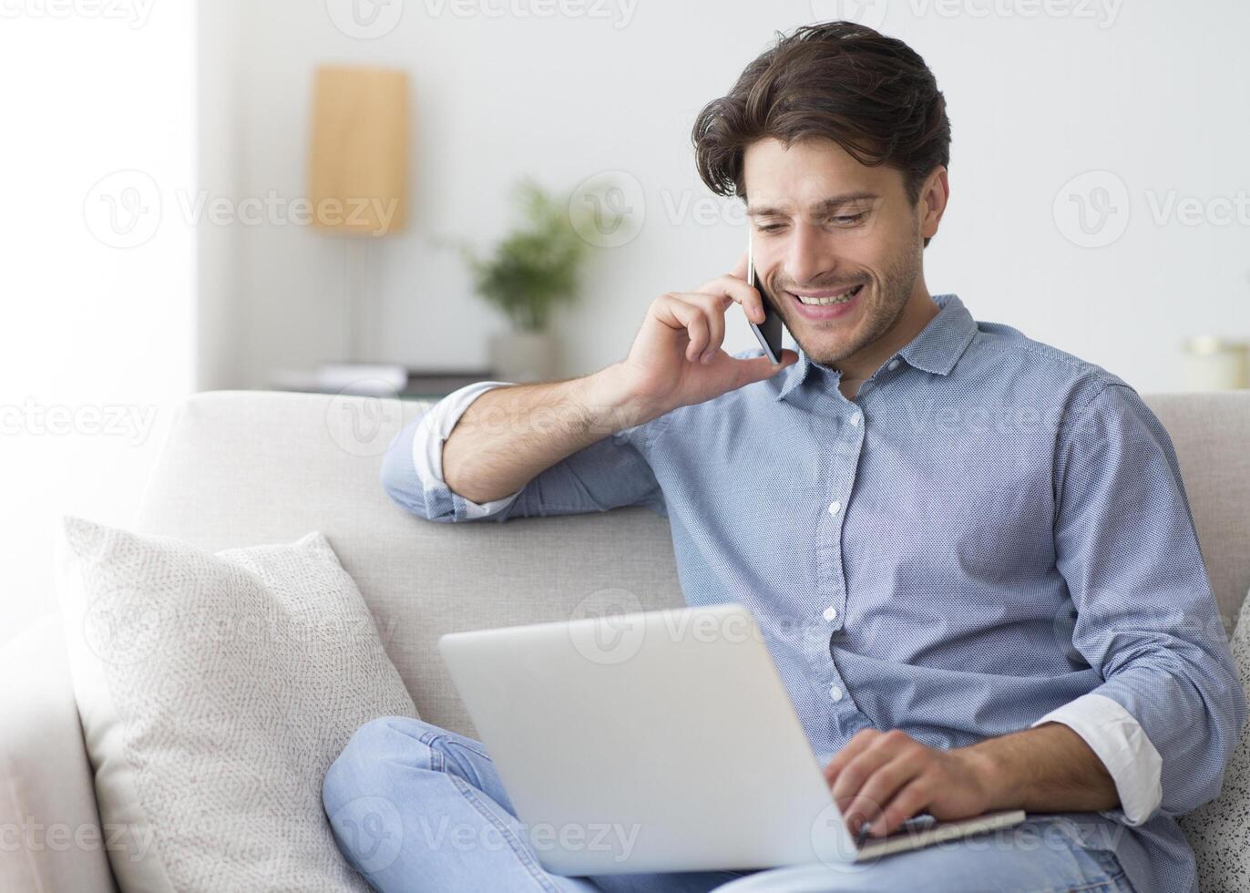 Always in touch. Successful entrepreneur talking on cellphone working at laptop sitting on sofa at home. Empty space photo